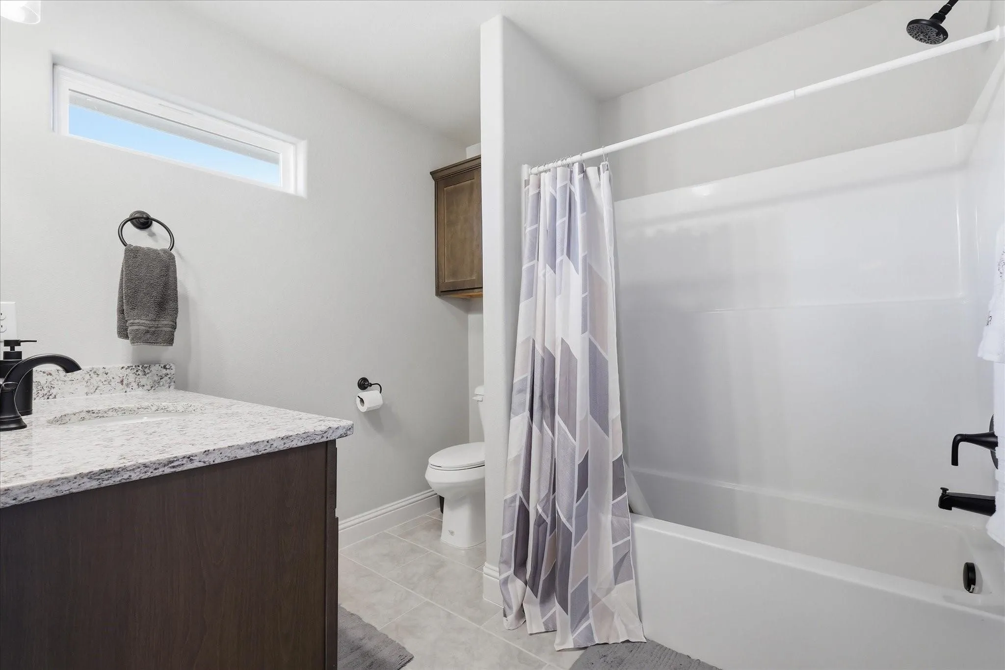 White bathtub with shower, dark wood-finish vanity, light-toned stone countertop, rectangular undermount sink, and matte black fixtures