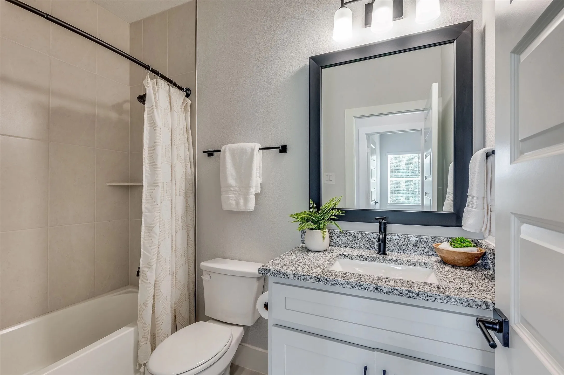 White paneled door, black-framed mirror, granite countertop, white vanity with black hardware, and a tiled shower surround