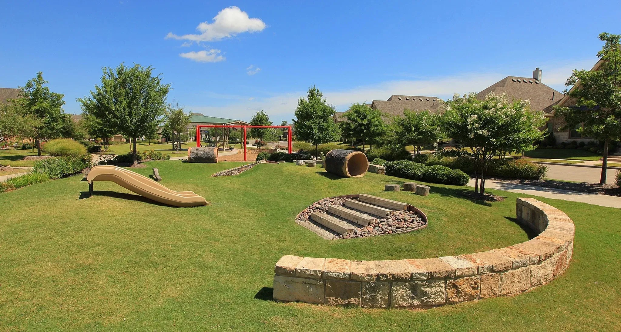 Expansive green space featuring a natural play area with a curved stone retaining wall, an integrated slide, and a tunnel structure