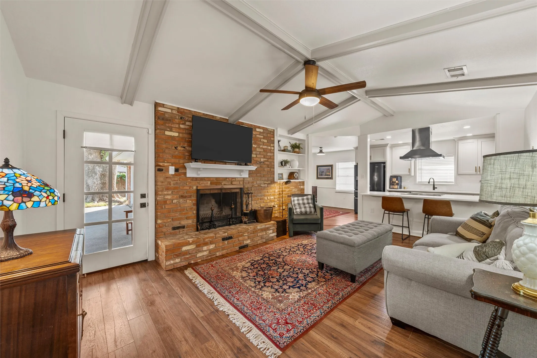 Vaulted ceiling with exposed beams, brick fireplace with mantel, wood-finish flooring, recessed lighting, and built-in shelving