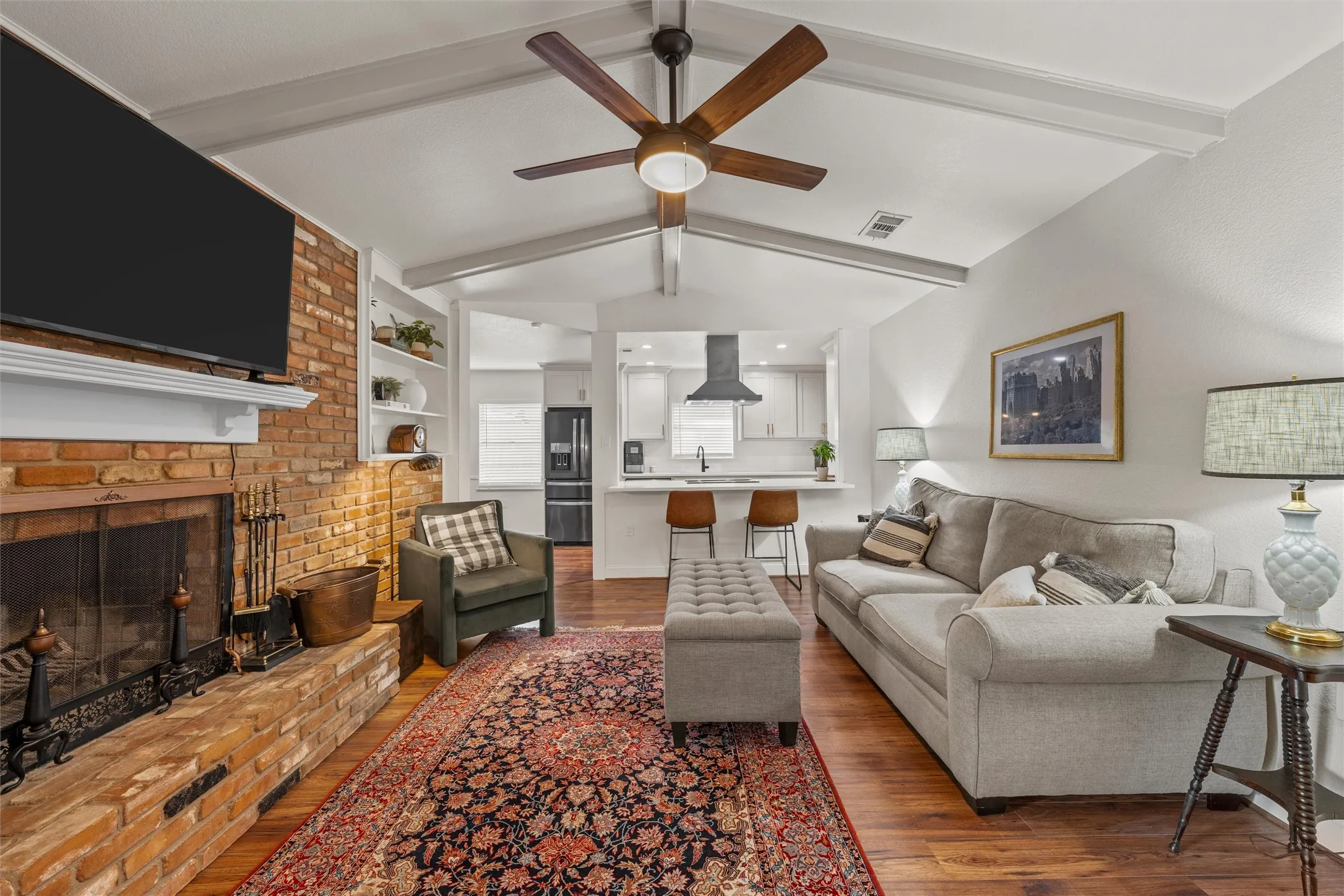 Vaulted ceiling with exposed beams, brick fireplace with mantel, wood-finish flooring, built-in shelving, and a modern ceiling fan