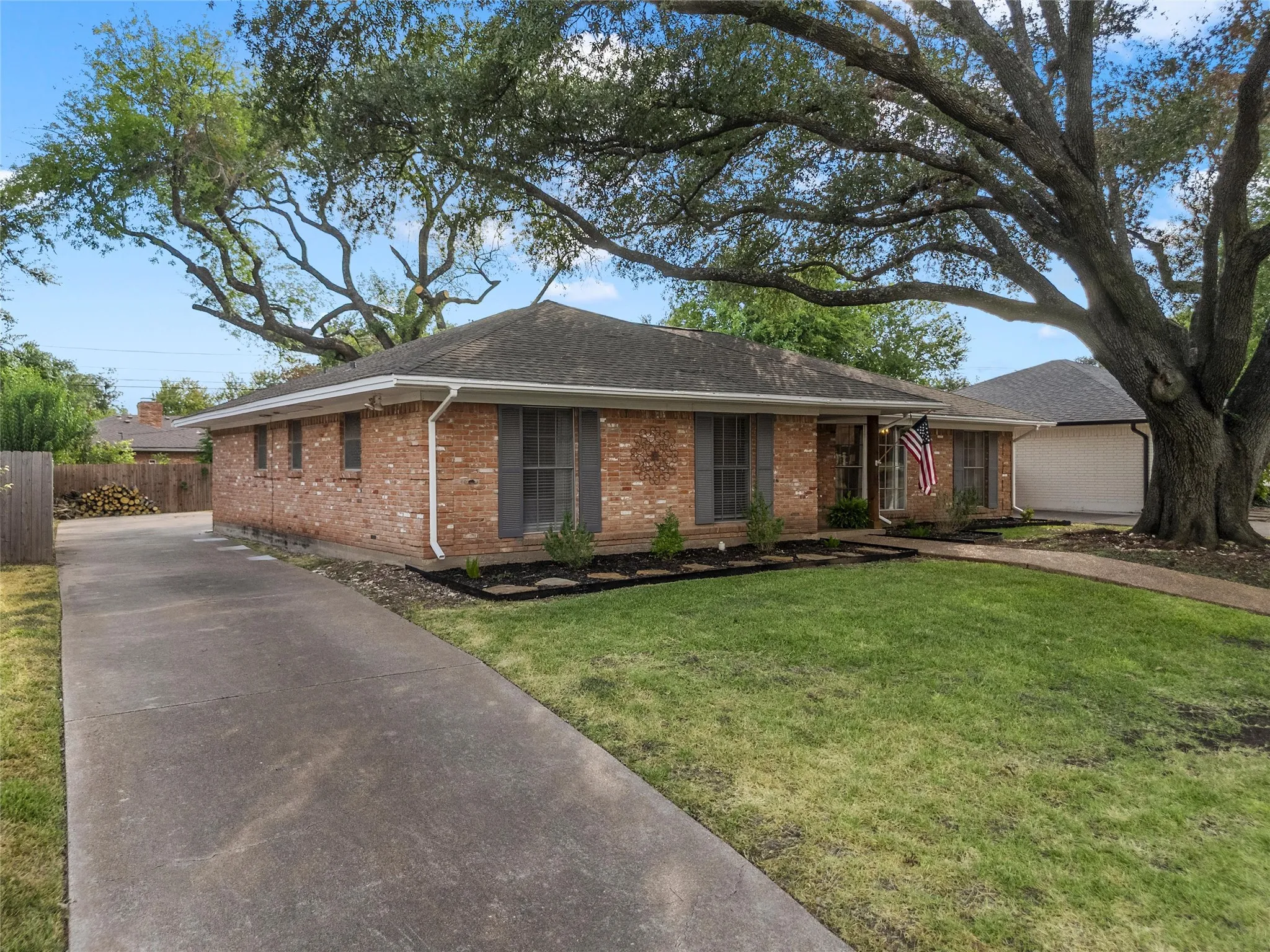 Brick exterior with dark gray shutters, mature front yard tree, established landscaping, and an extended concrete driveway