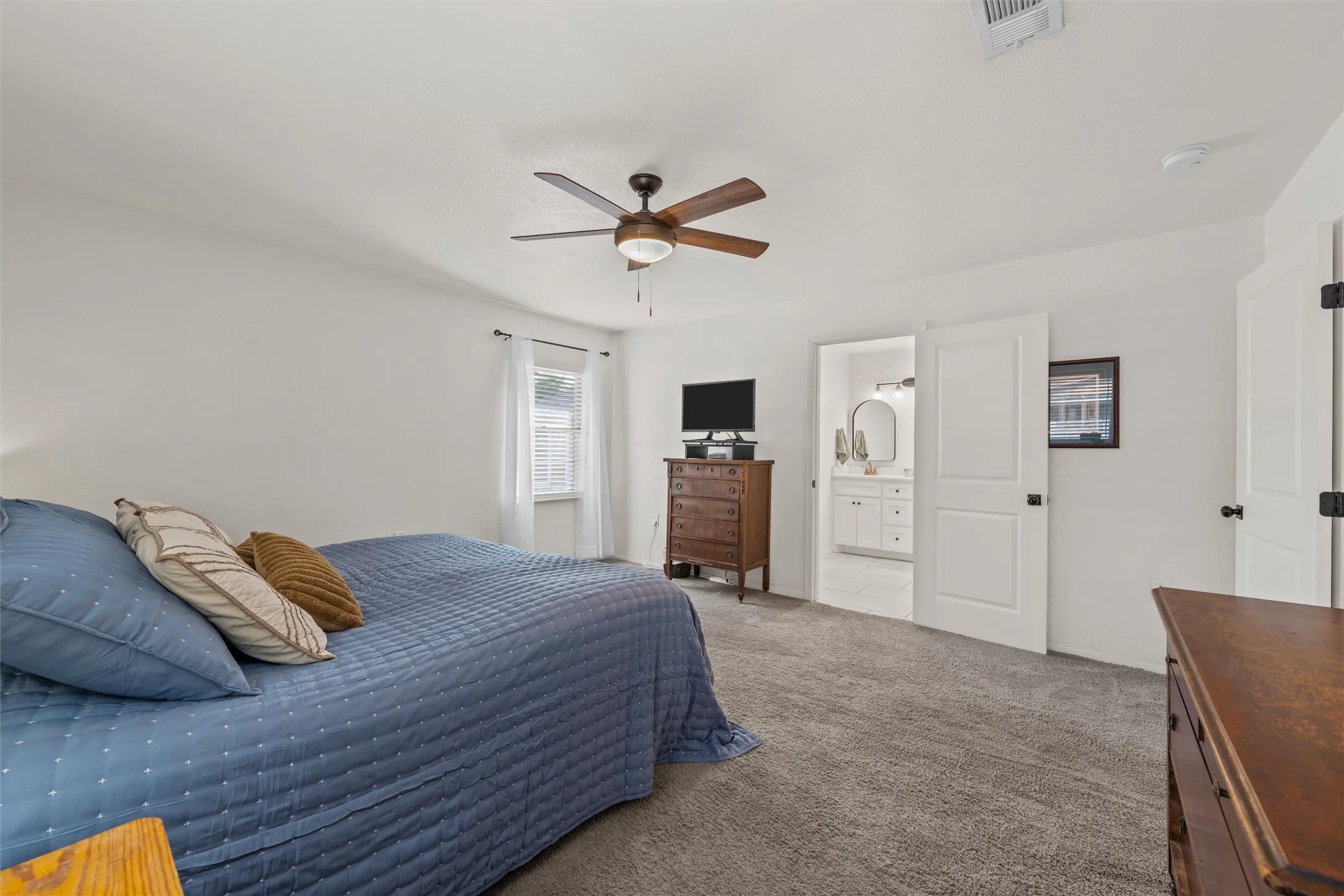 Plush carpet flooring, a ceiling fan with integrated lighting, and a white paneled door leading to a bathroom with a white vanity