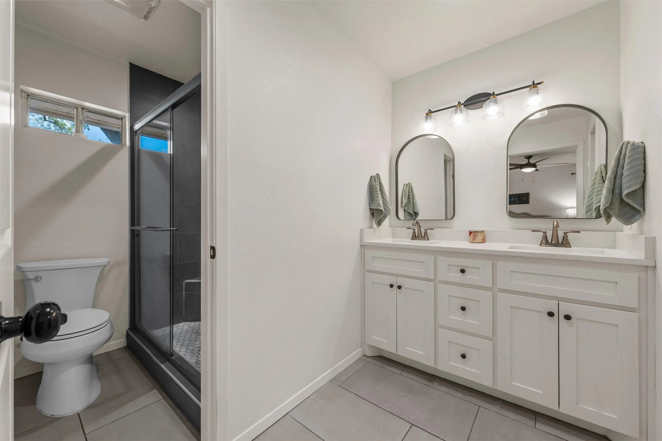 Dual vanity with white cabinetry and integrated sinks, a walk-in shower with dark-tiled walls, a toilet, and gray tile flooring