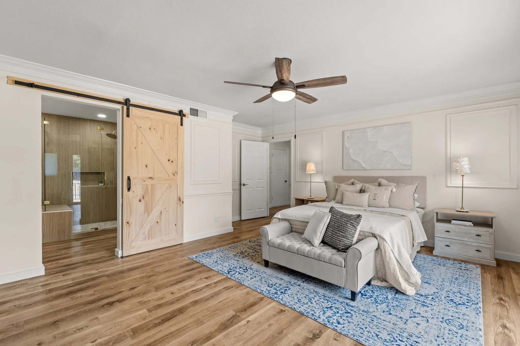 Wood-finish flooring throughout, ceiling fan with integrated lighting, crown molding, and a barn door entry to the adjacent bath featuring a glass-enclosed shower