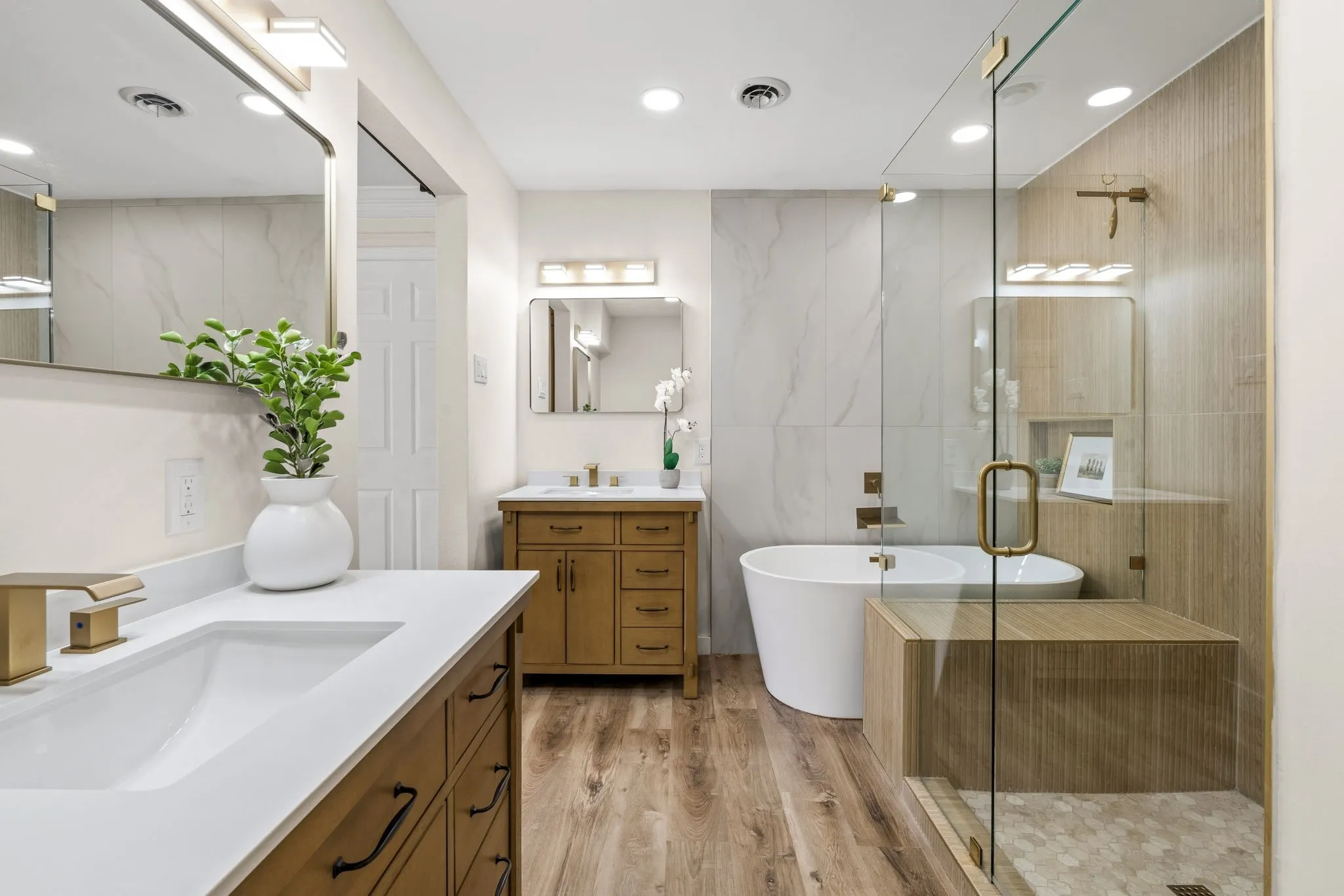 Dual vanities with white countertops and gold-tone fixtures, a freestanding soaking tub, a glass-enclosed shower with a built-in bench, and wood-finish flooring