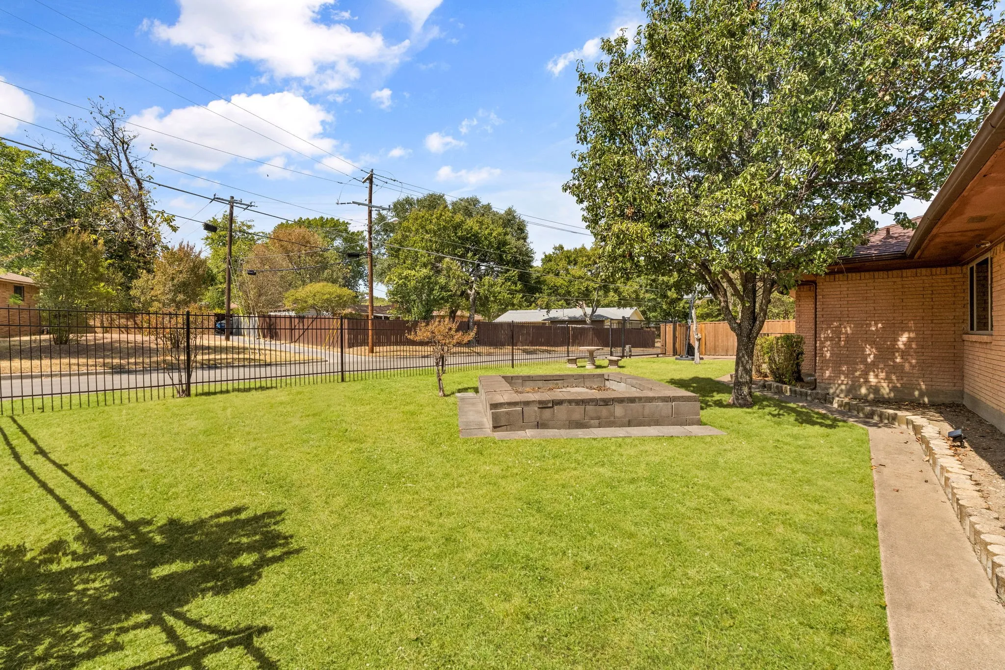Expansive yard featuring a concrete fire pit, mature shade tree, brick exterior, and a wrought iron fence