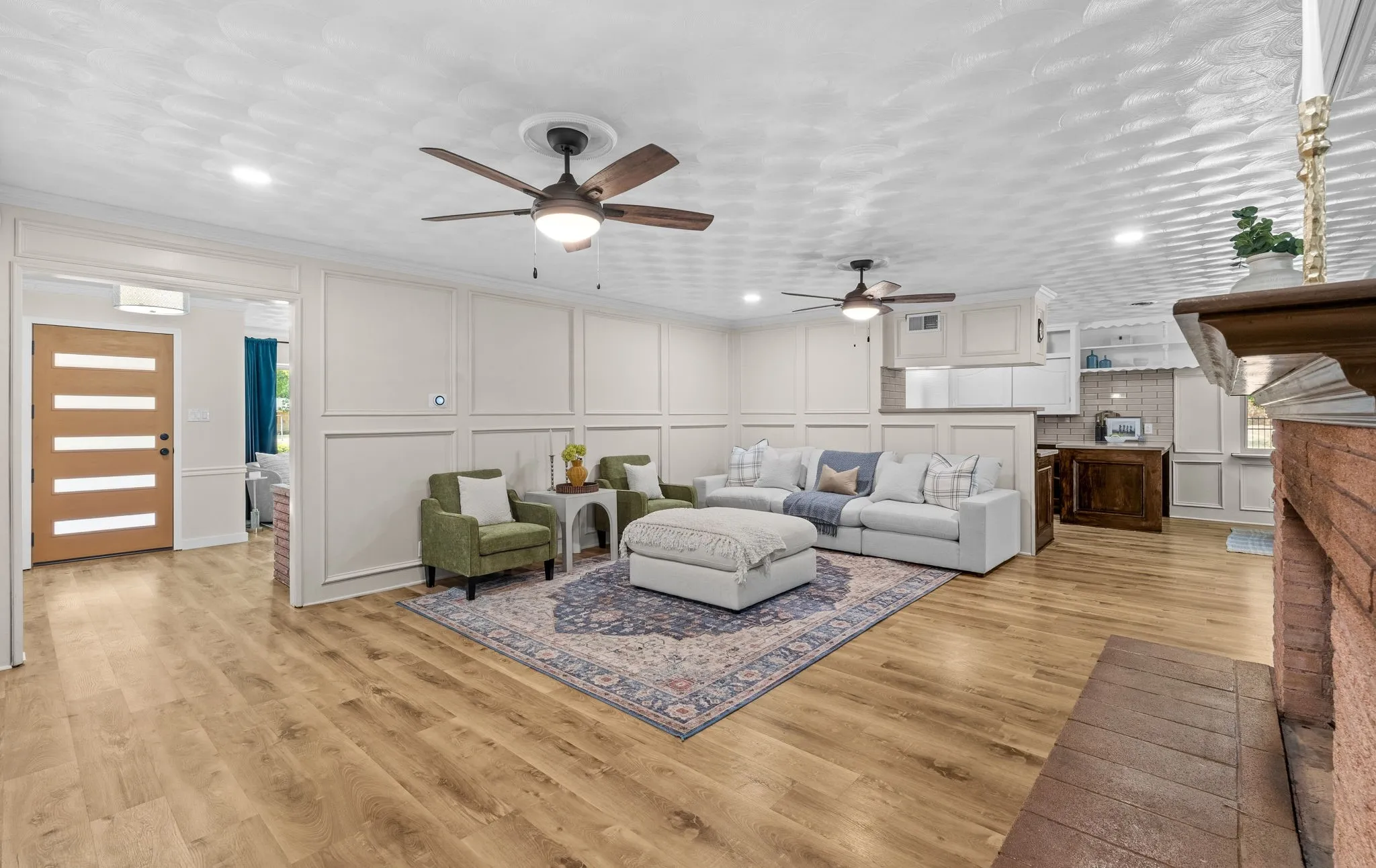 Wood-finish flooring throughout, two ceiling fans with integrated lighting, recessed lighting, white wainscoting paneling, and a brick fireplace