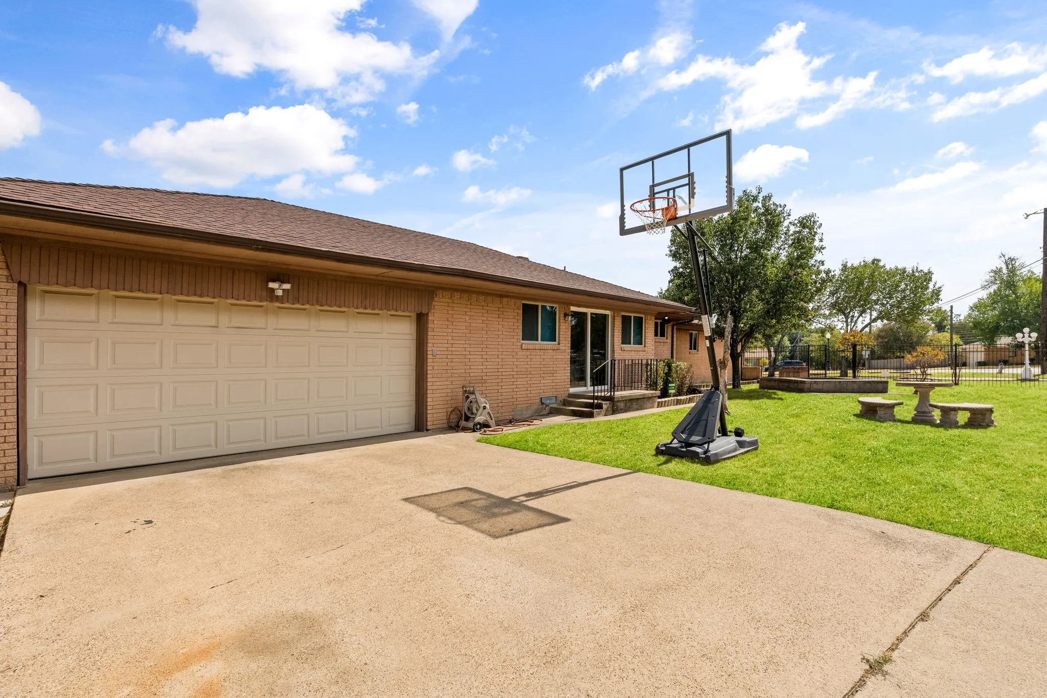 Brick exterior featuring an attached garage, concrete driveway, and a spacious lawn with a stone picnic table