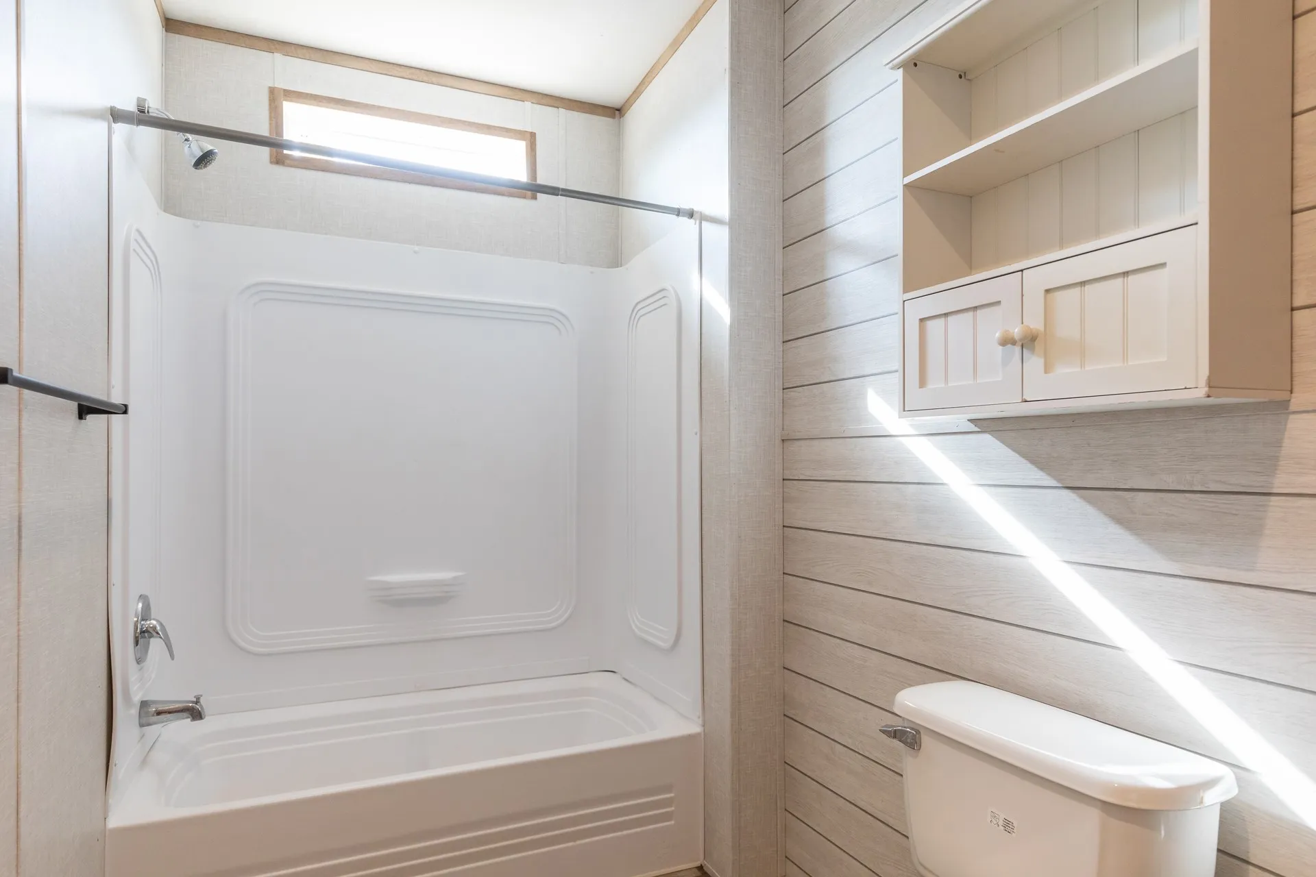 White bathtub and shower combination with chrome fixtures, an overhead transom window, plank-style wall paneling, and an integrated wall cabinet with open shelving