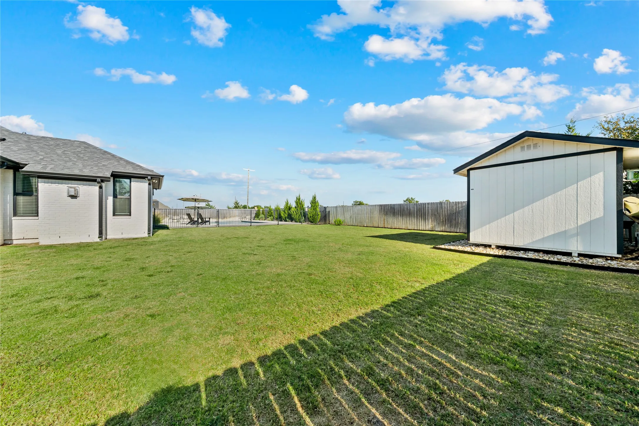 Expansive green space featuring a white siding shed, a wood fence, and a white stucco exterior building