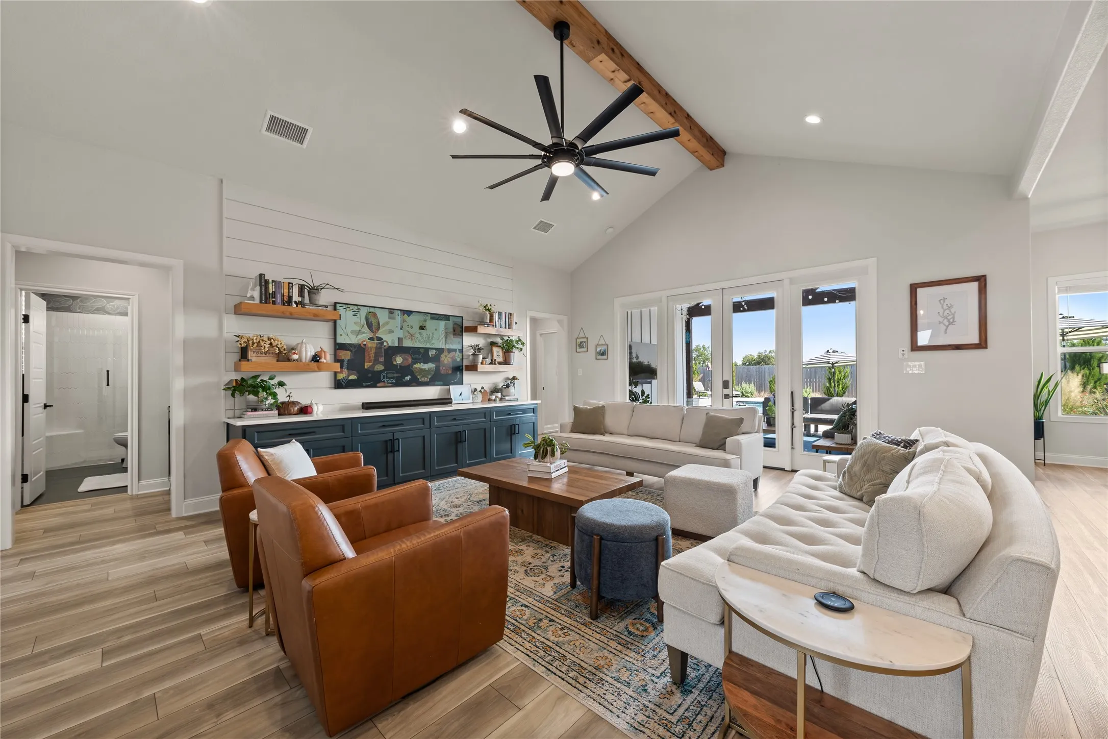 Vaulted ceiling with exposed wood beam, wood-finish flooring, built-in entertainment center with open shelving, large ceiling fan, and sliding glass doors