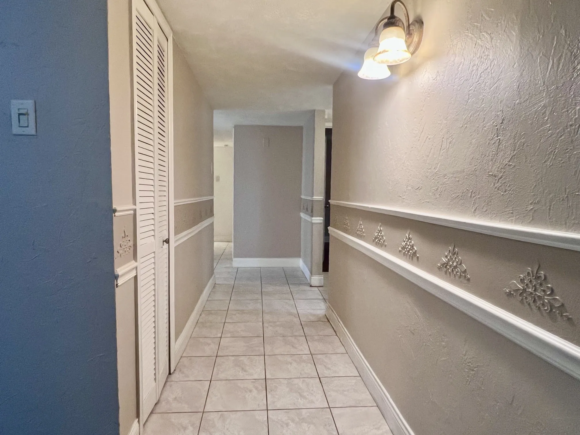 Hallway featuring tile flooring, textured walls, chair rail molding with decorative appliques, and a built-in storage closet with louvered bifold doors