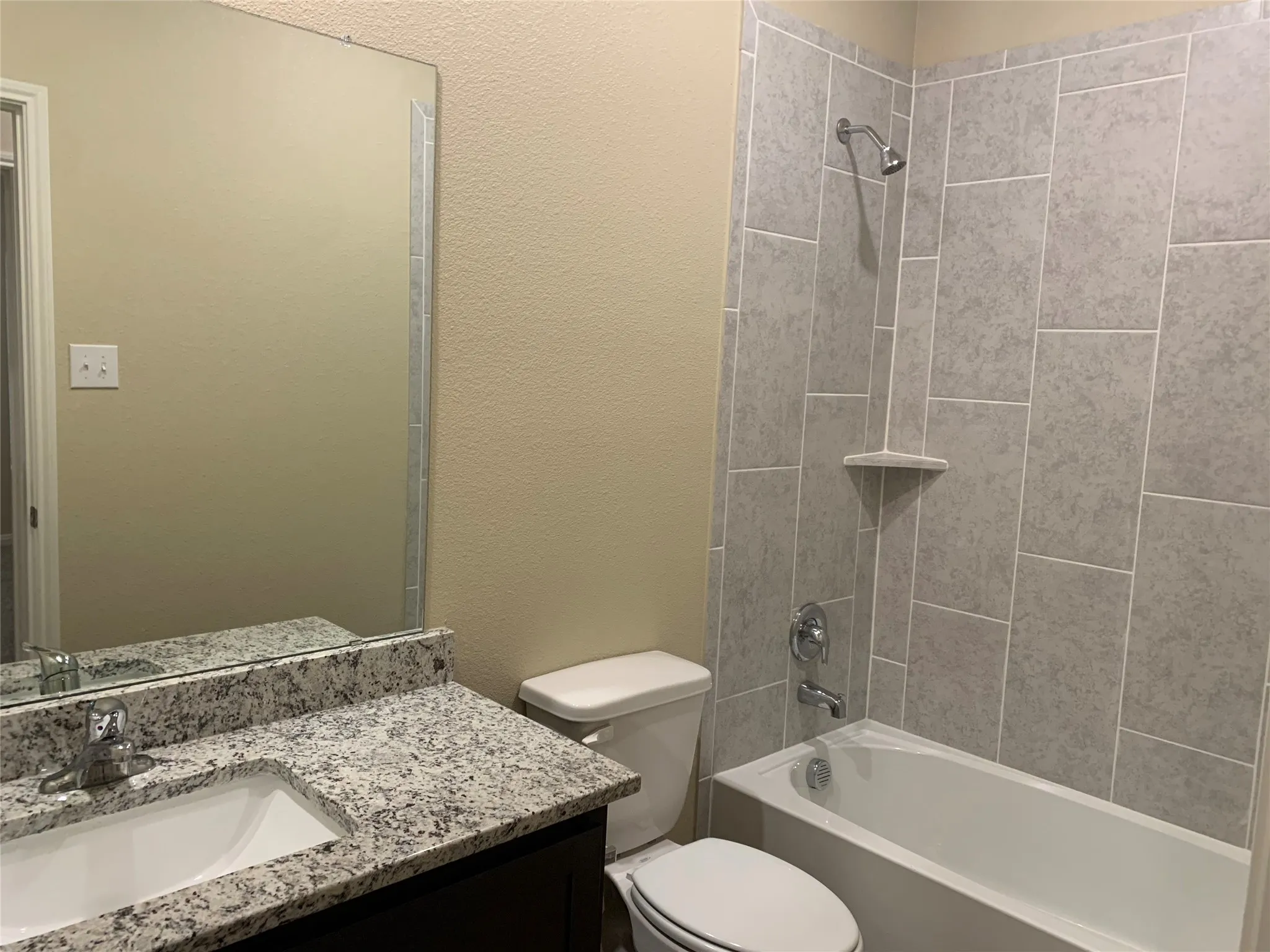 Vanity with built-in sink and stone-finish countertop, mirror with decorative trim, and shower-tub combination with tiled surround and built-in shelf