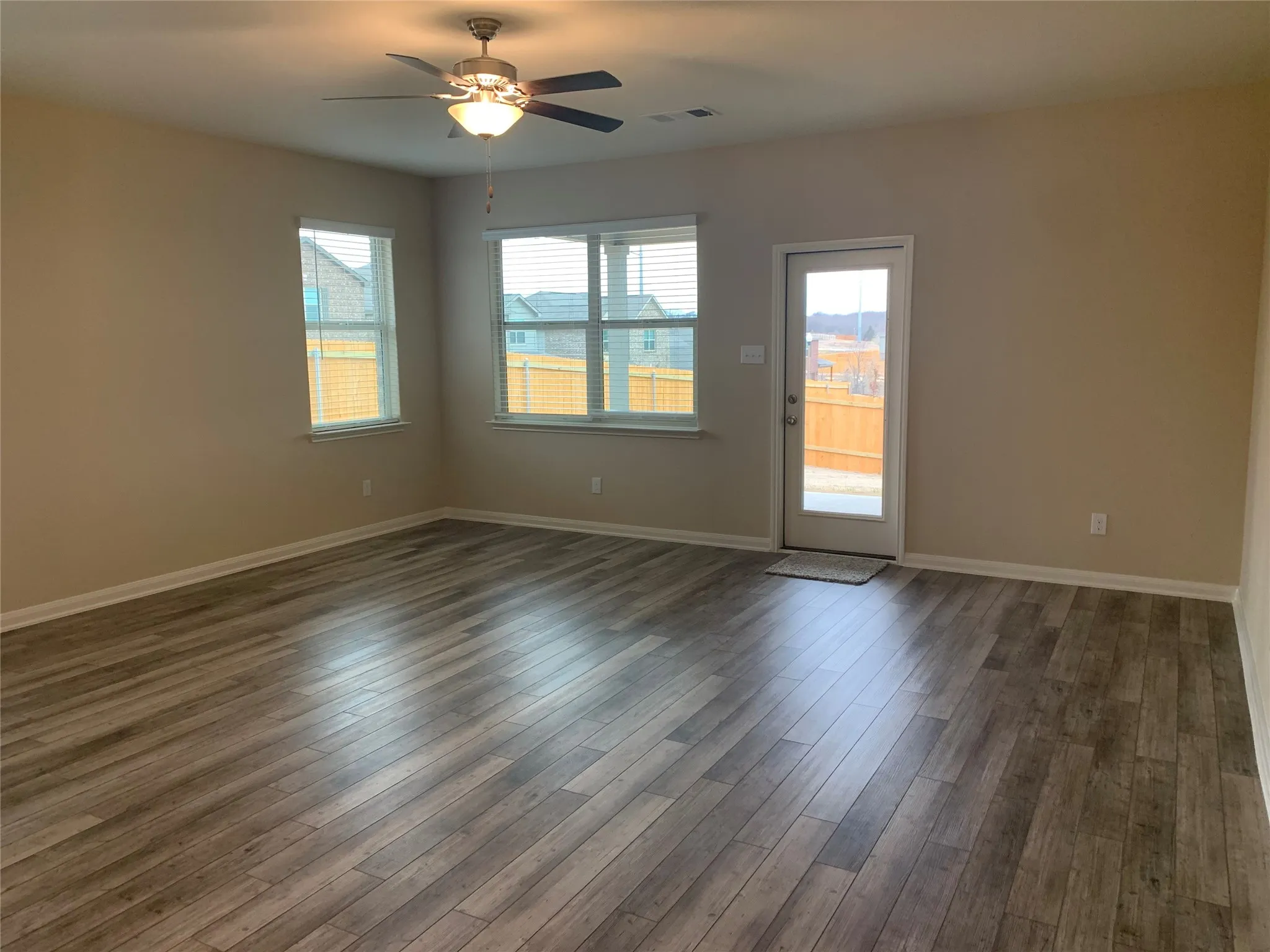 Expansive room featuring wood-finish flooring, neutral-toned walls, a ceiling fan with integrated lighting, multiple windows, and a glass-paneled exterior door