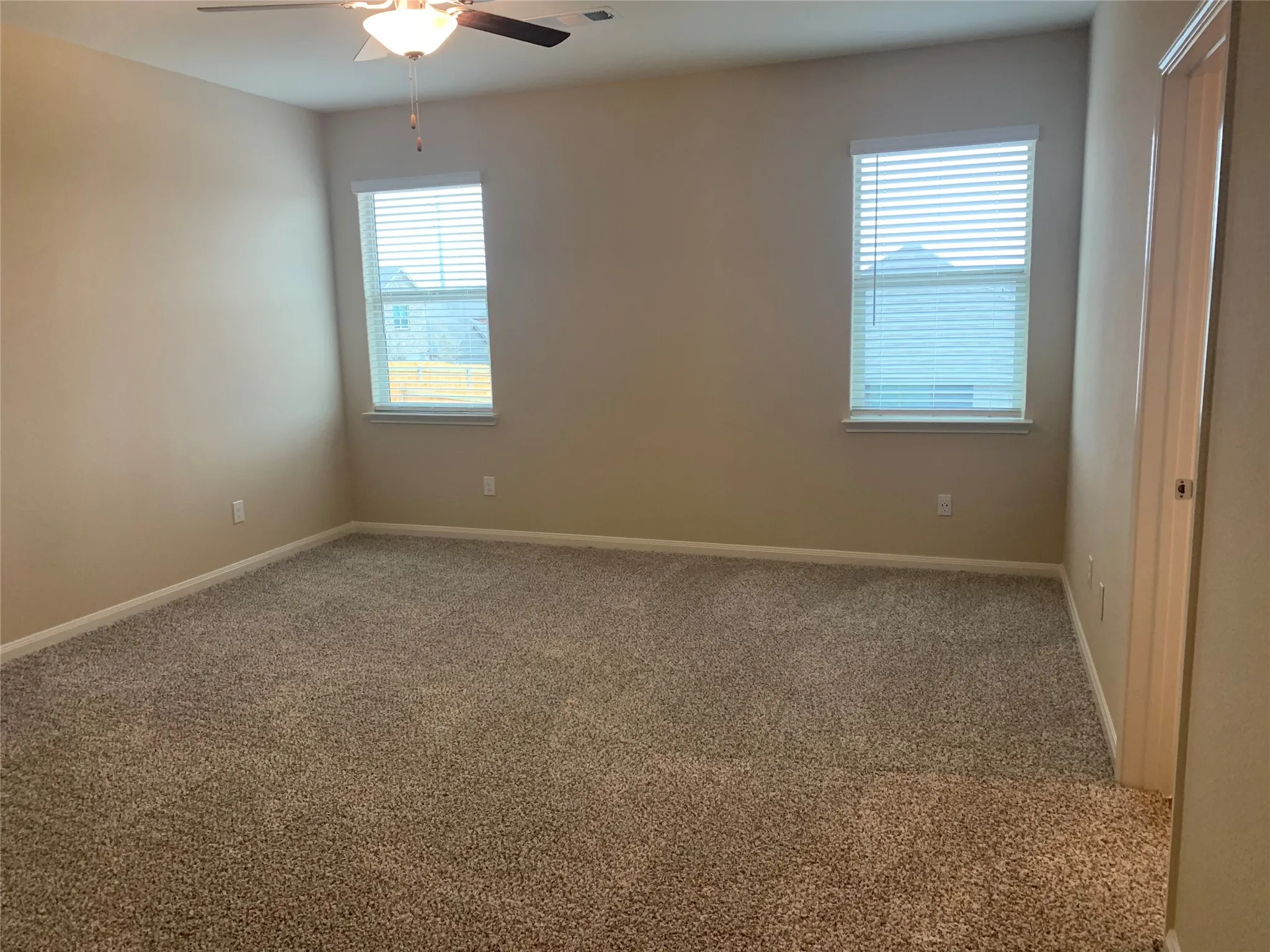 Neutral wall color, two windows with blinds, ceiling fan with integrated lighting, and plush carpeting