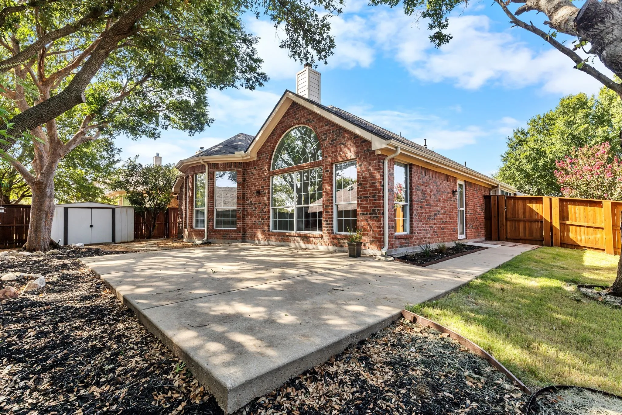 Brick exterior with an arched window, multiple large windows, a concrete patio, a wooden fence, and a storage shed
