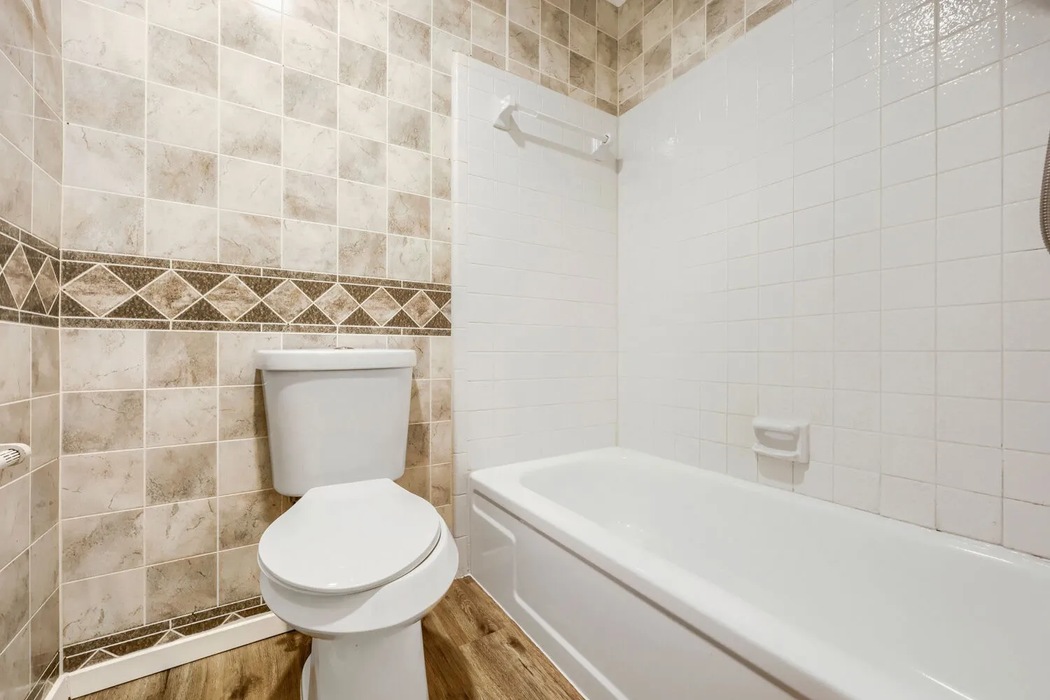 Full-size bathtub with white tile surround, wood-finish flooring, and wall-mounted toilet