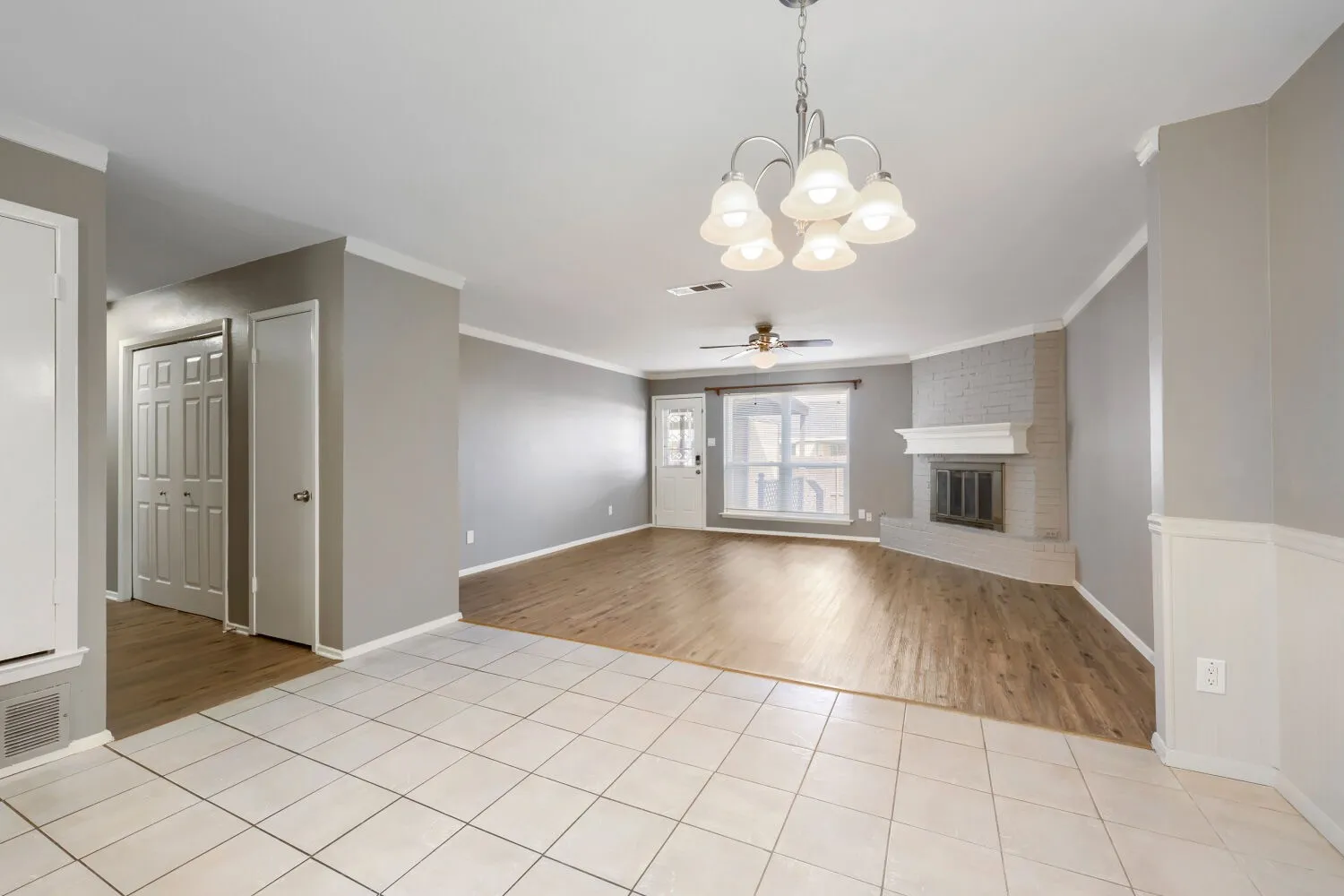 Open-concept living area featuring mixed tile and wood-finish flooring, a painted brick fireplace with a mantle, and an overhead chandelier