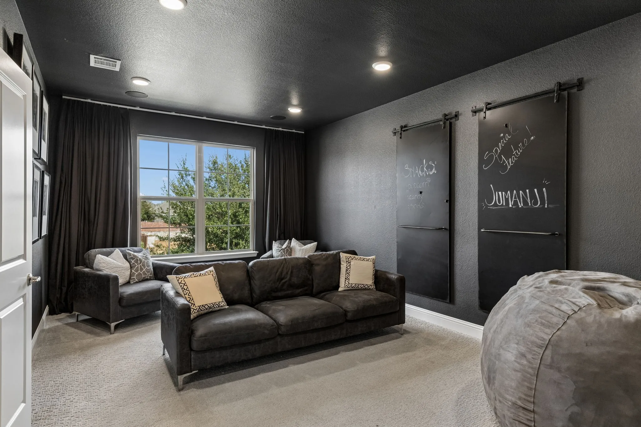 Sophisticated media room creates an inviting destination for movie nights and entertaining, featuring rich charcoal walls and ceiling, recessed lighting, comfortable seating area, and custom chalkboard-style barn doors that add a distinctive designer touch.