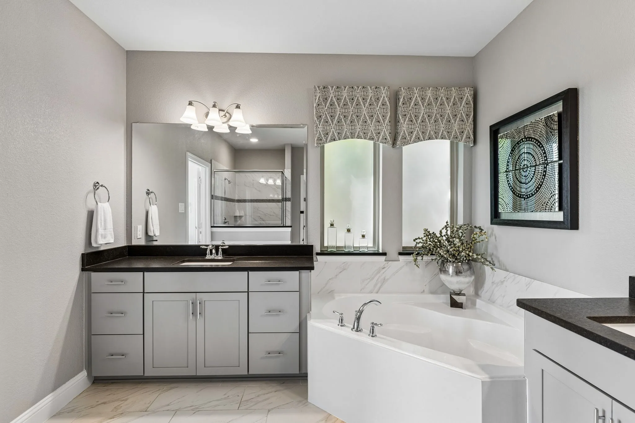 This elegant primary bath showcases an expansive vanity with generous storage, sophisticated dark countertops, oversized mirror, refined fixtures, and a calming neutral palette.