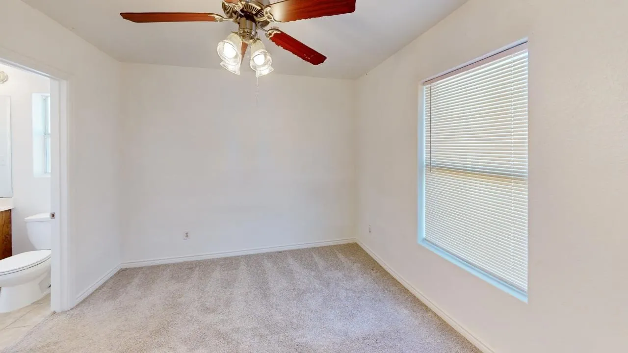 Carpeted room featuring a ceiling fan with light fixture, a window with blinds, and an adjacent bathroom with a toilet and vanity
