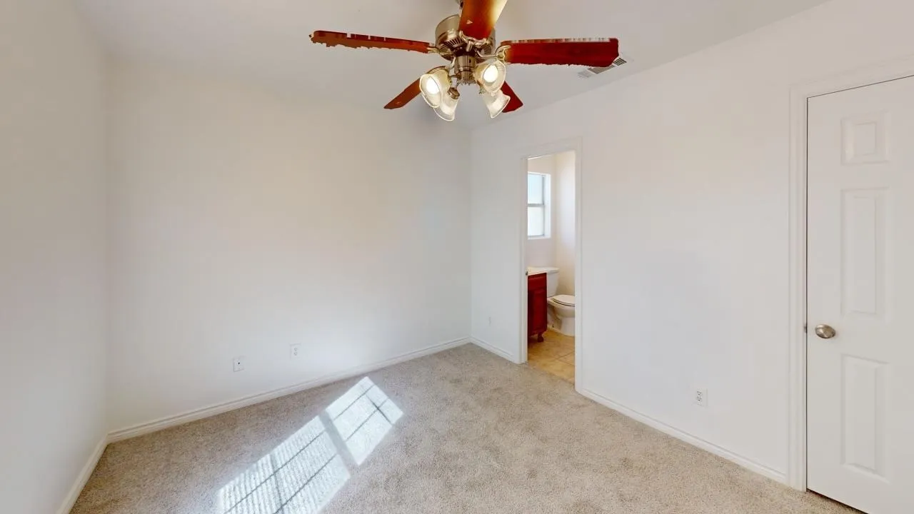 Carpeted flooring, white interior walls, overhead ceiling fan with lighting, white interior door, and a partially visible bathroom with a dark wood-finish vanity