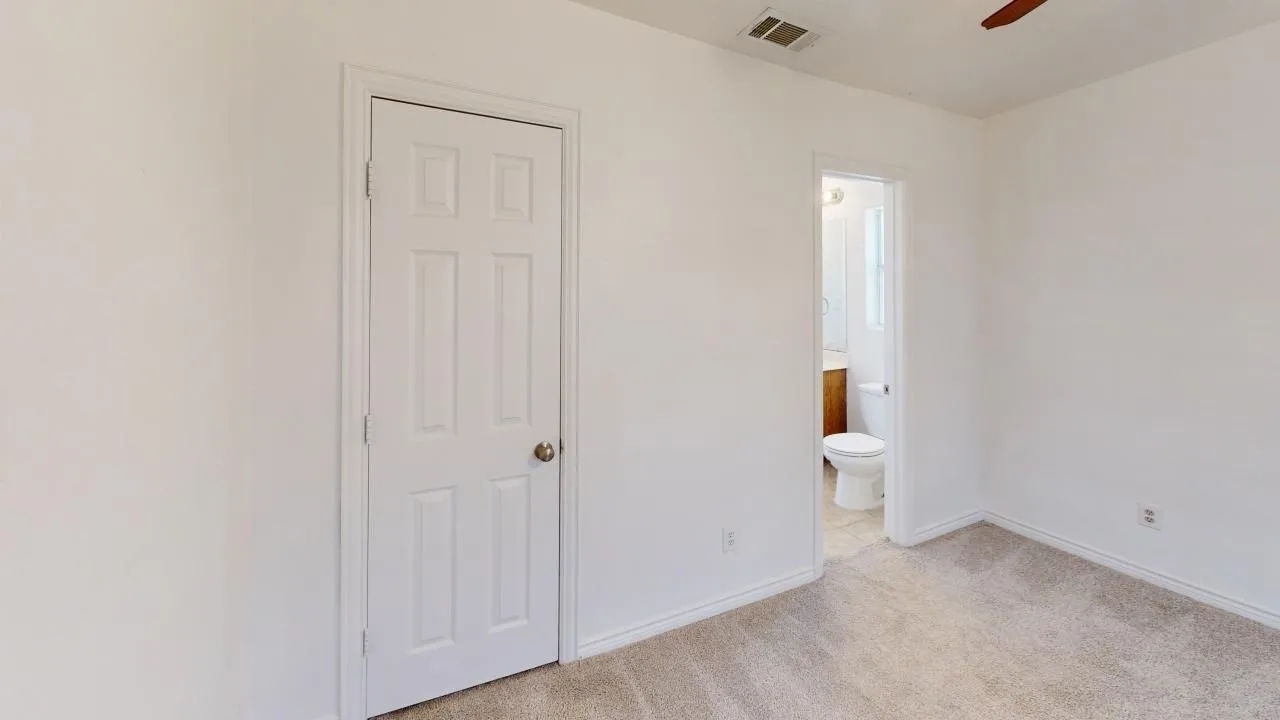 Carpeted room with white walls, a six-panel interior door, and a partially visible bathroom with a toilet and wood-finish vanity