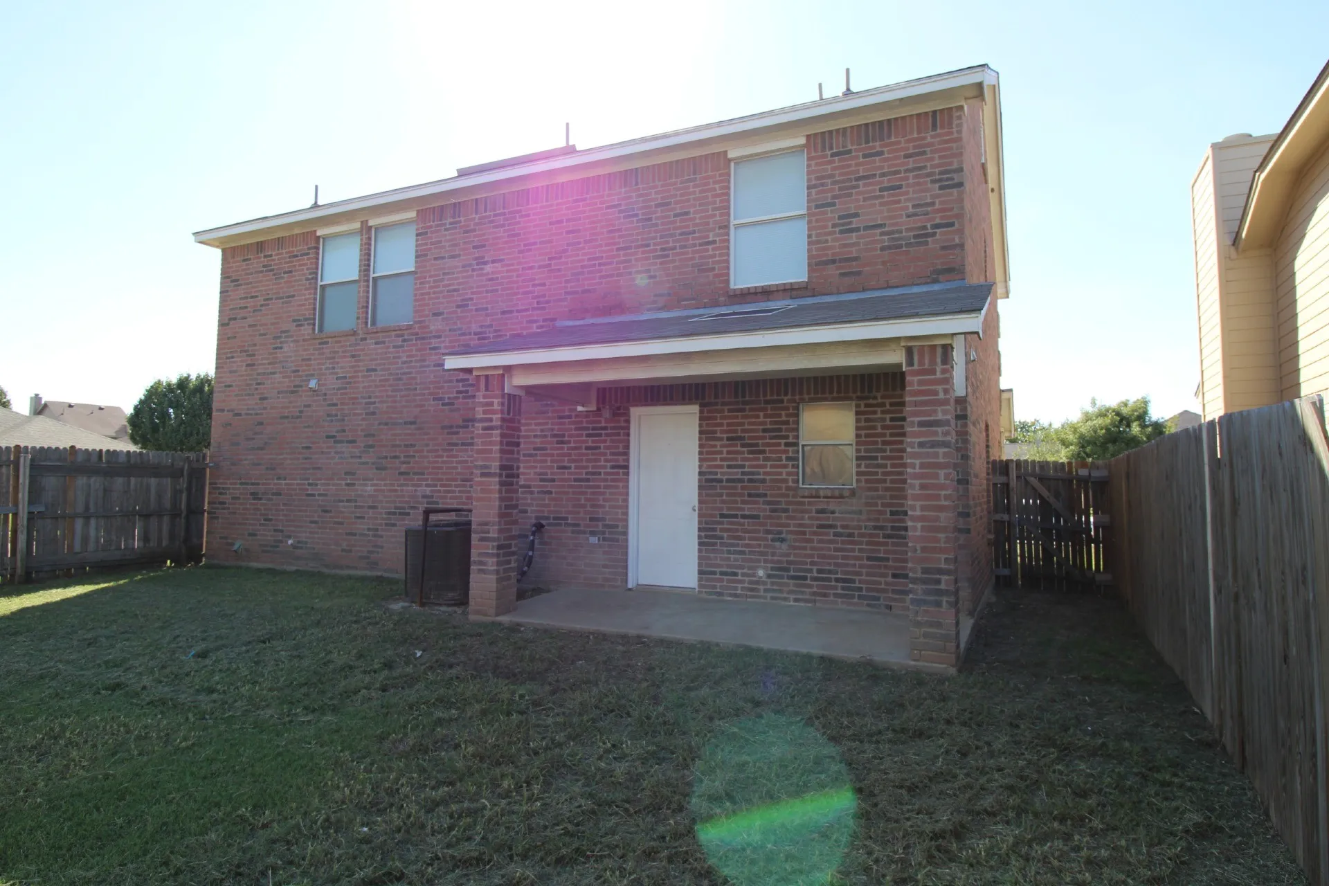 Brick Colonial home featuring a two-story elevation with multiple windows, a covered rear entry, and a spacious yard with mature landscaping and wooden perimeter fencing