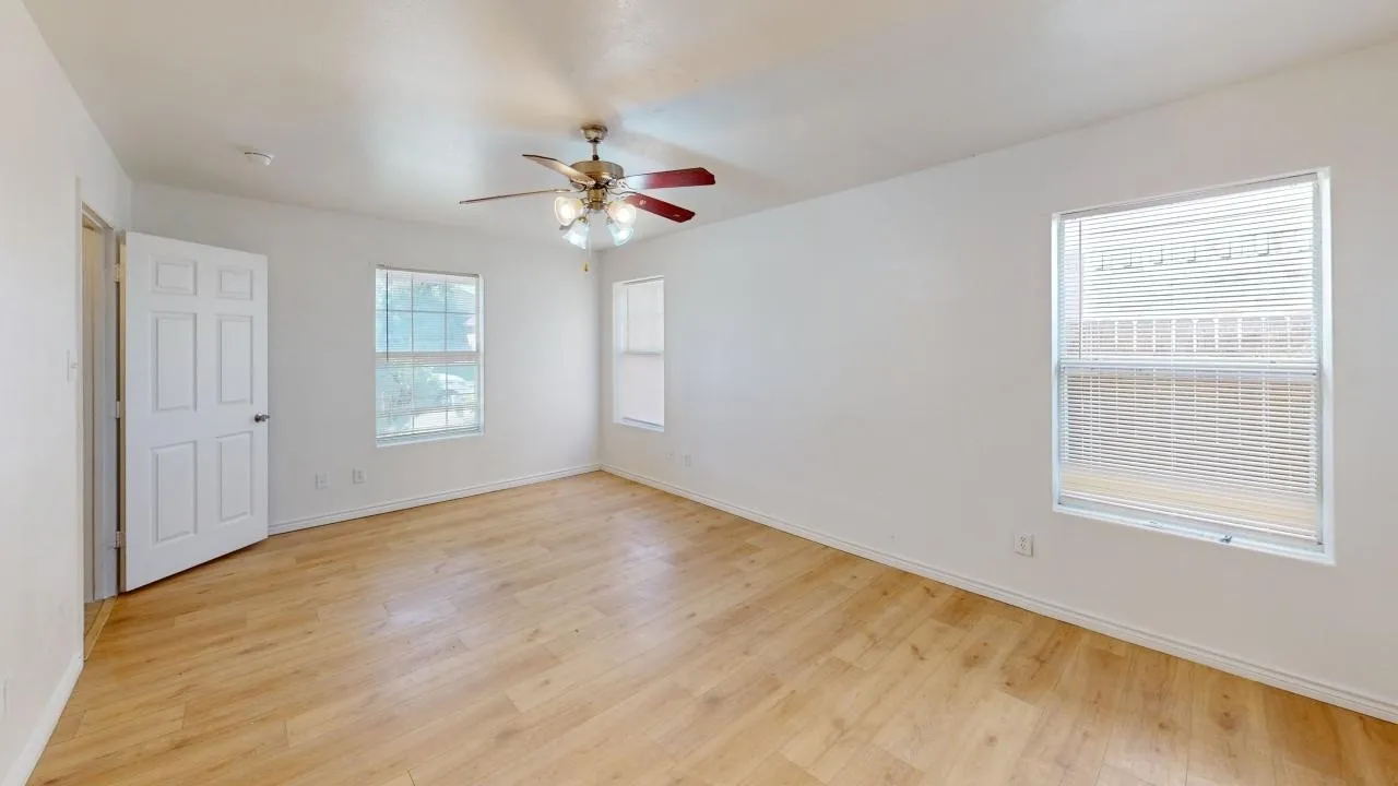 Bright, open room showcasing gleaming wood-finish flooring throughout, abundant natural light from multiple windows with operable blinds, ceiling fan with integrated lighting fixture, and white six-panel entry door