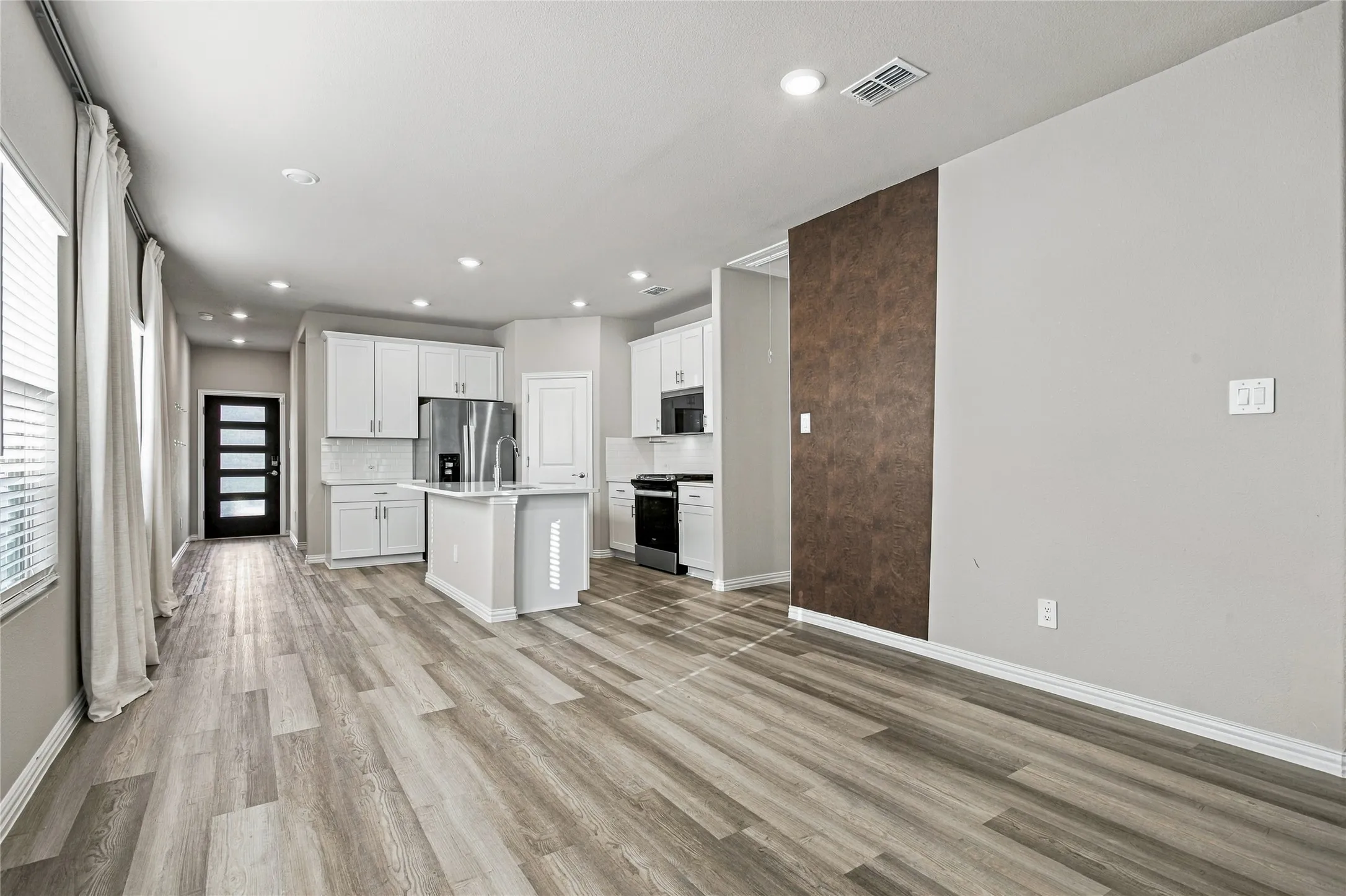 Wood-finish flooring throughout, recessed ceiling lighting, kitchen island with built-in sink, white cabinetry with integrated appliances, and a modern entry door