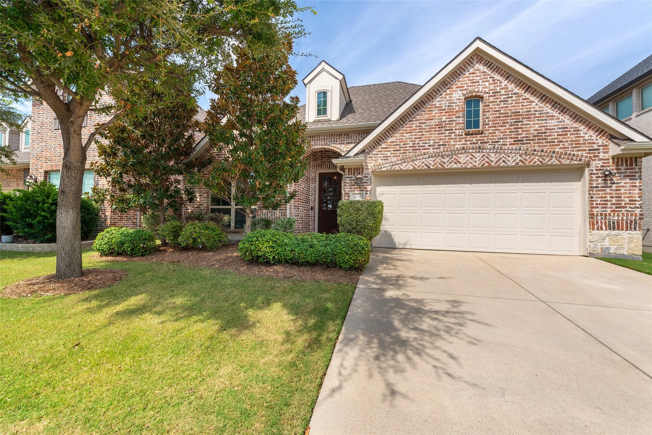 Brick facade with stone accents, an attached garage, a dormer window, mature trees, and landscaped shrubbery