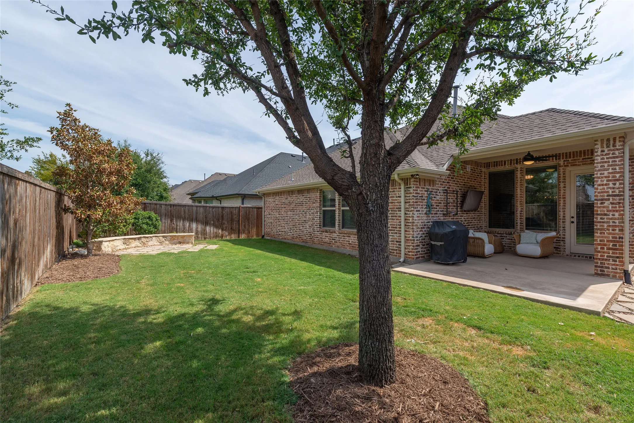 Expansive backyard oasis featuring mature shade trees, manicured lawn, and a covered patio with brick detailing