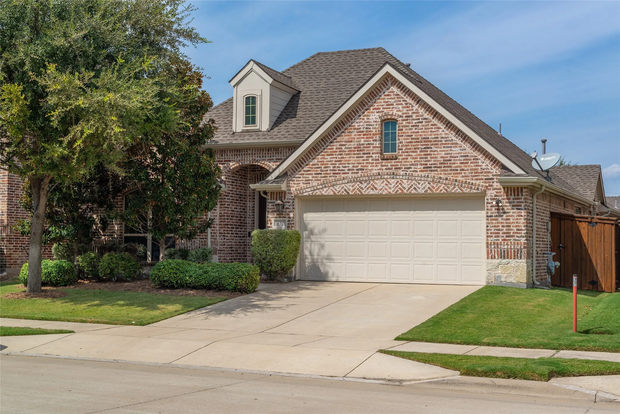 Brick facade with stone accents, dormer window, attached garage, concrete driveway, and manicured landscaping