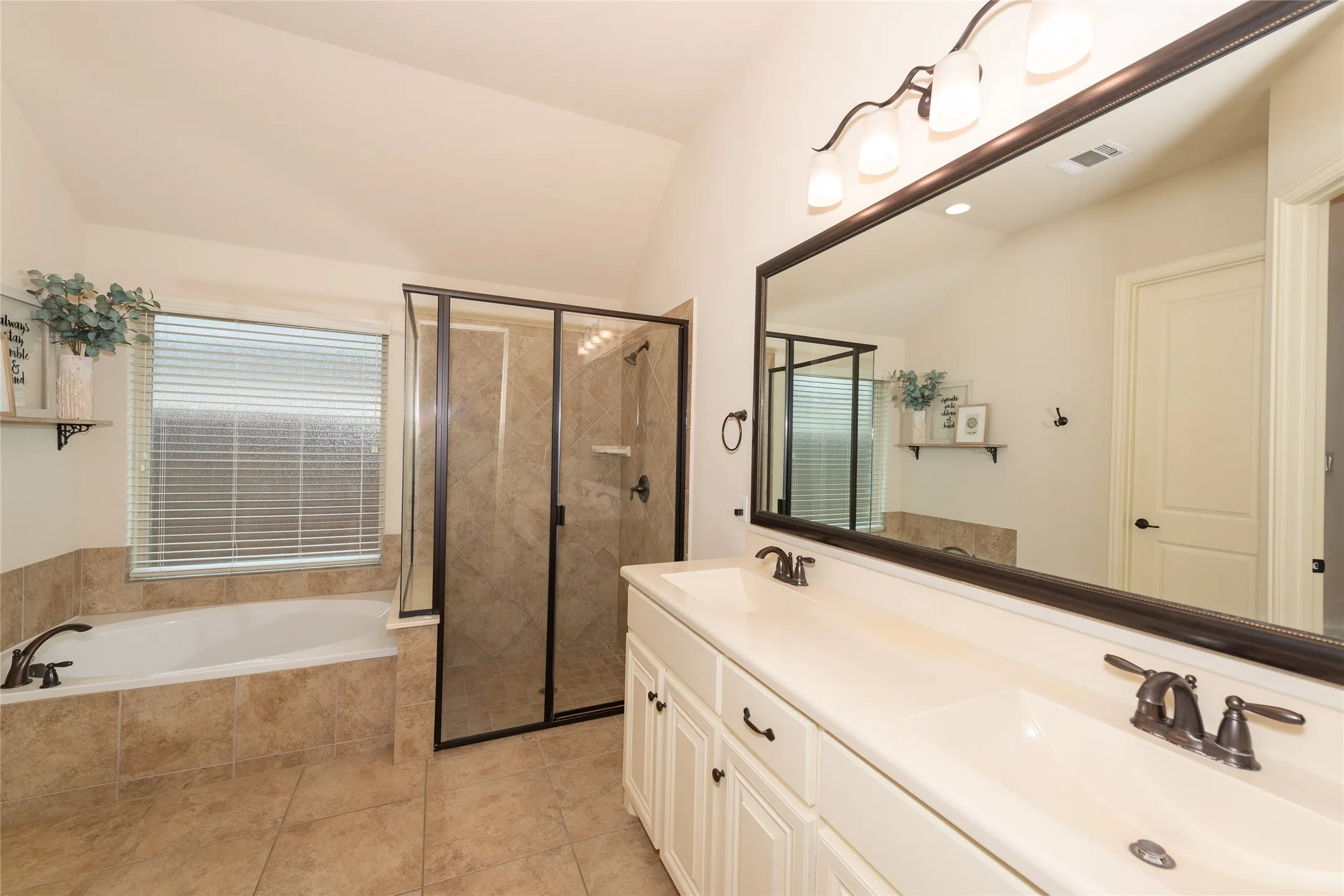 Spacious bathroom featuring a dual vanity with integrated sinks, a large framed mirror, a separate soaking tub, and a glass-enclosed shower with tiled walls