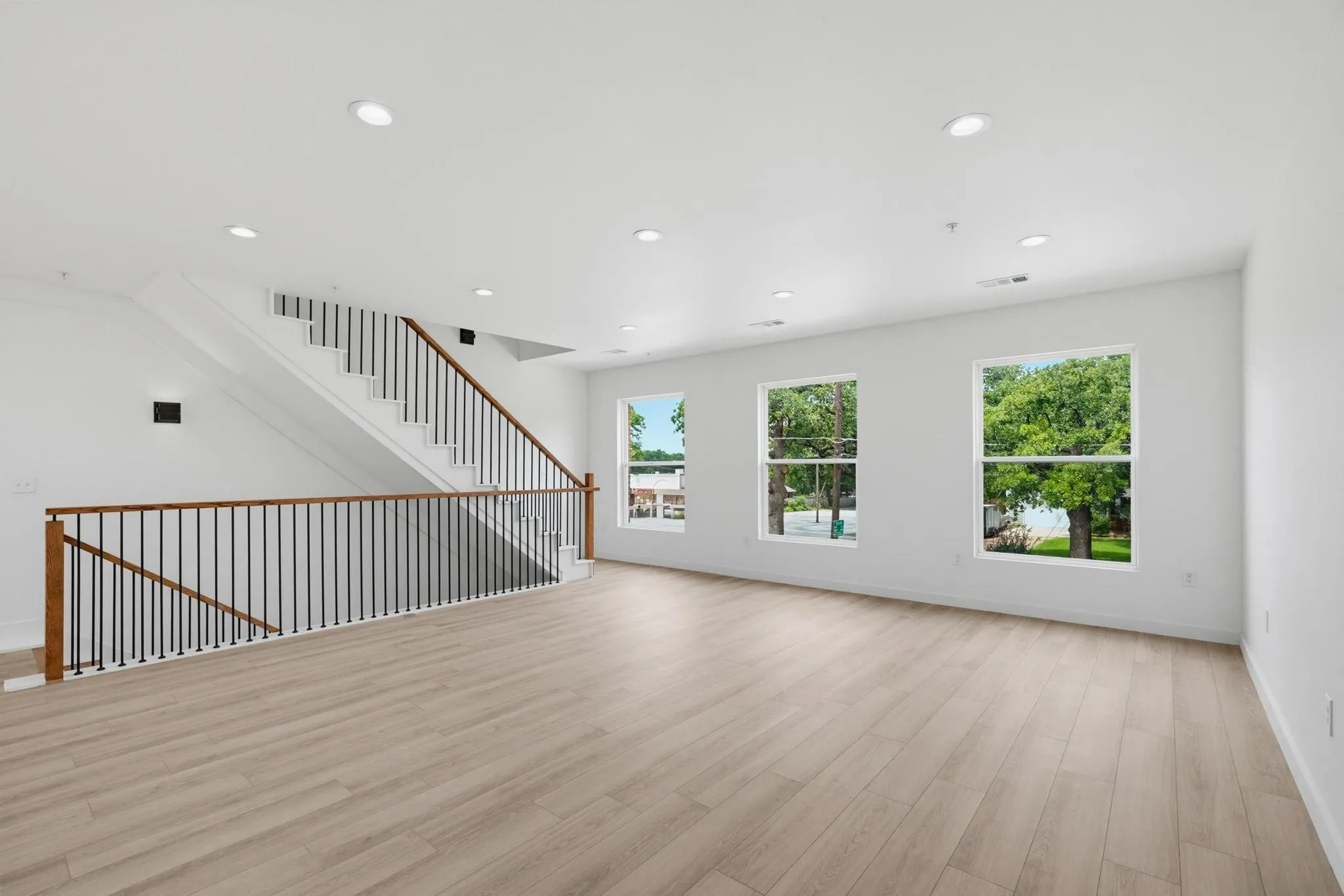 Light wood-finish flooring, white walls, recessed ceiling lighting, three windows, and a staircase with a wood handrail and black metal balusters