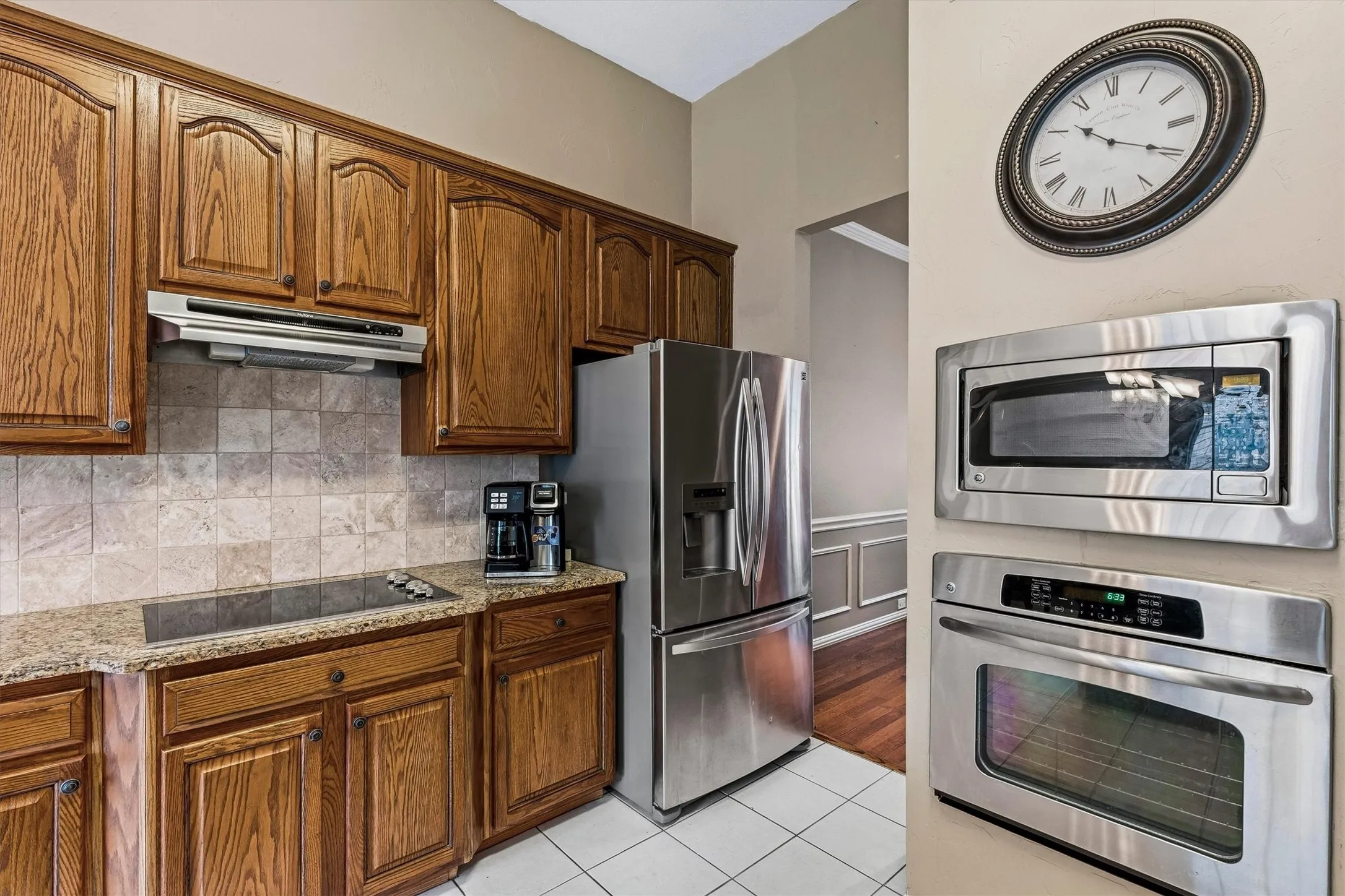 Wood cabinetry, granite-finish countertop, tile backsplash, built-in cooktop, and stainless steel appliances