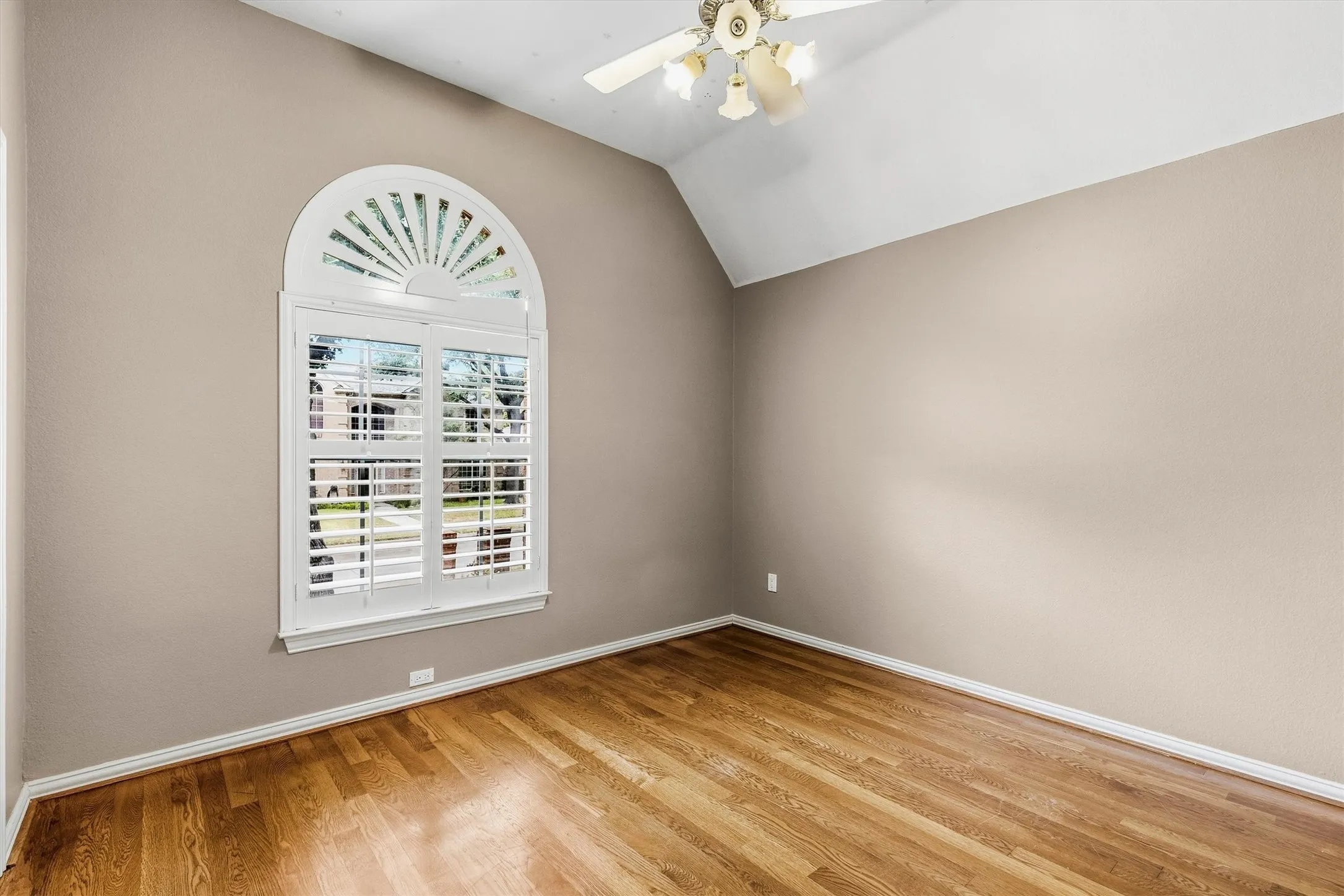 Bright upper-level bedroom featuring warm wood-finish flooring, neutral taupe walls, and an arched transom window with plantation shutters