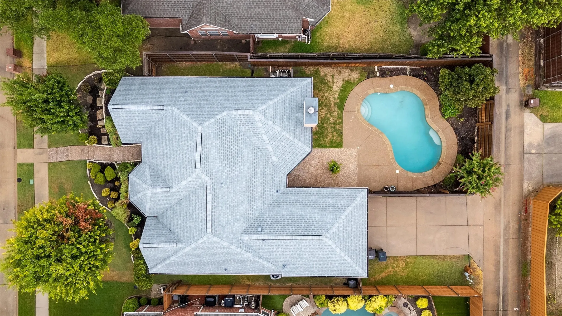 Aerial view showcasing a light-colored roof with geometric paneling, resort-style free-form swimming pool with sun deck, mature landscaping throughout the property, and expansive hardscaped entertaining areas adjacent to lush green spaces