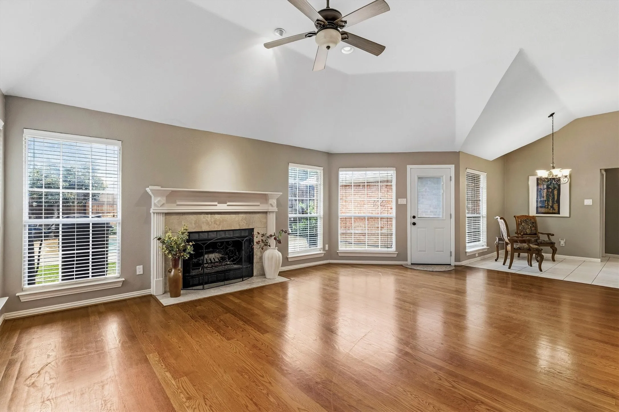 Spacious great room showcasing gleaming hardwood flooring, a traditional fireplace with white mantel and surround, vaulted ceiling with ceiling fan, and abundant natural light from multiple windows and glass doors