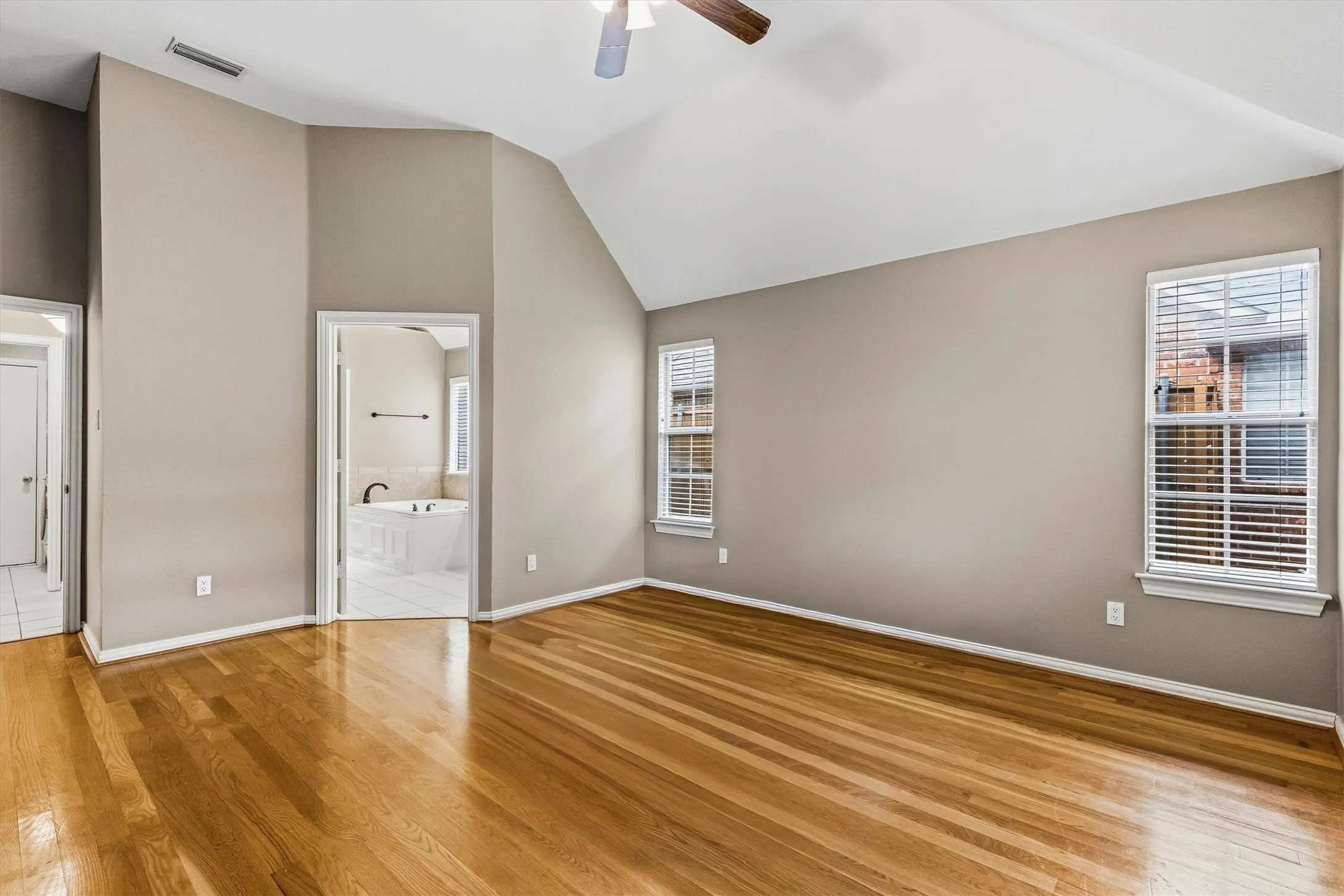 Vaulted ceiling, wood-finish flooring, two windows with blinds, ceiling fan, and access to an adjoining bathroom with a built-in tub