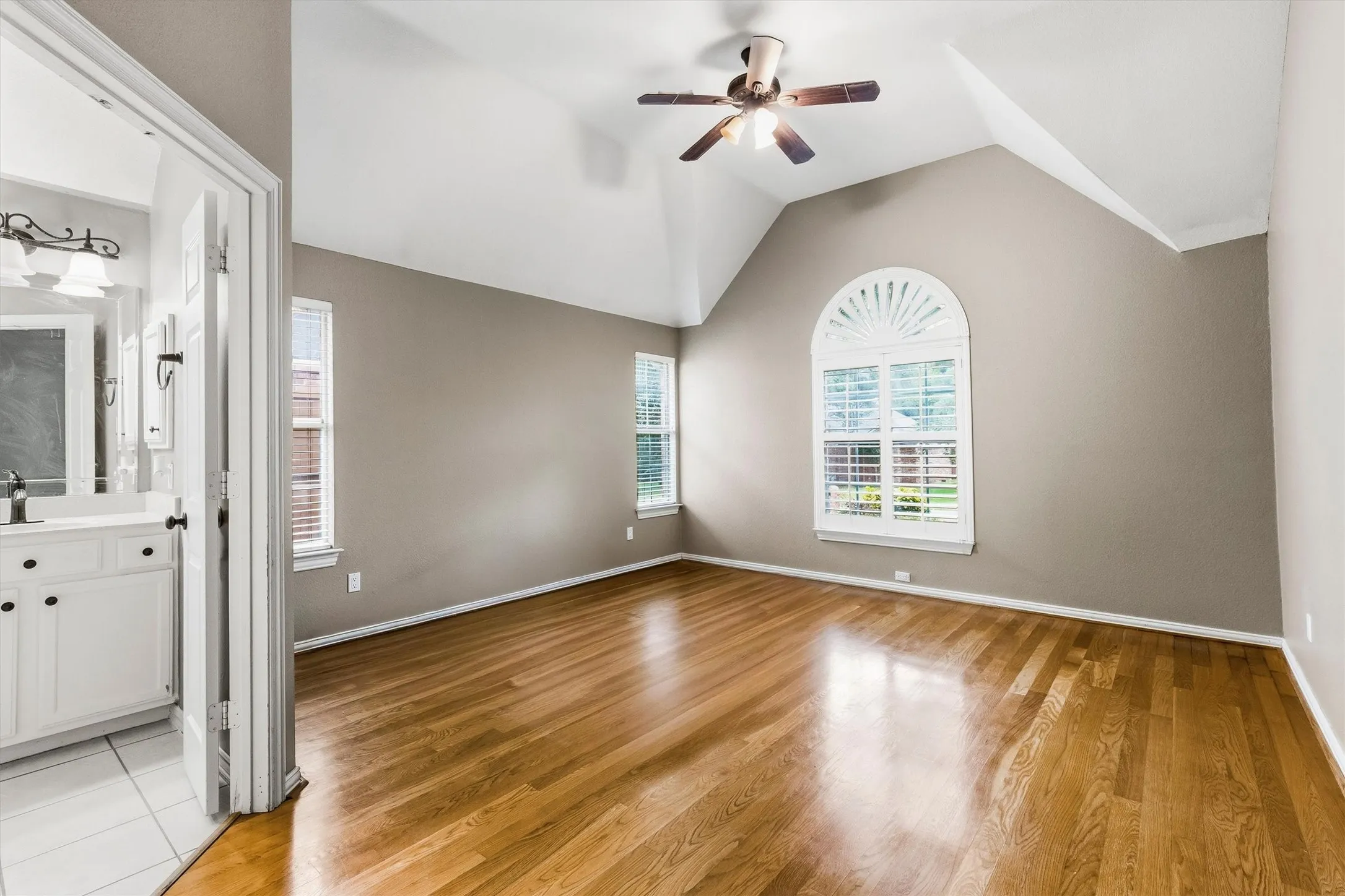 Vaulted ceiling, wood-finish flooring, ceiling fan, arched window with plantation shutters, and a vanity with a rectangular mirror