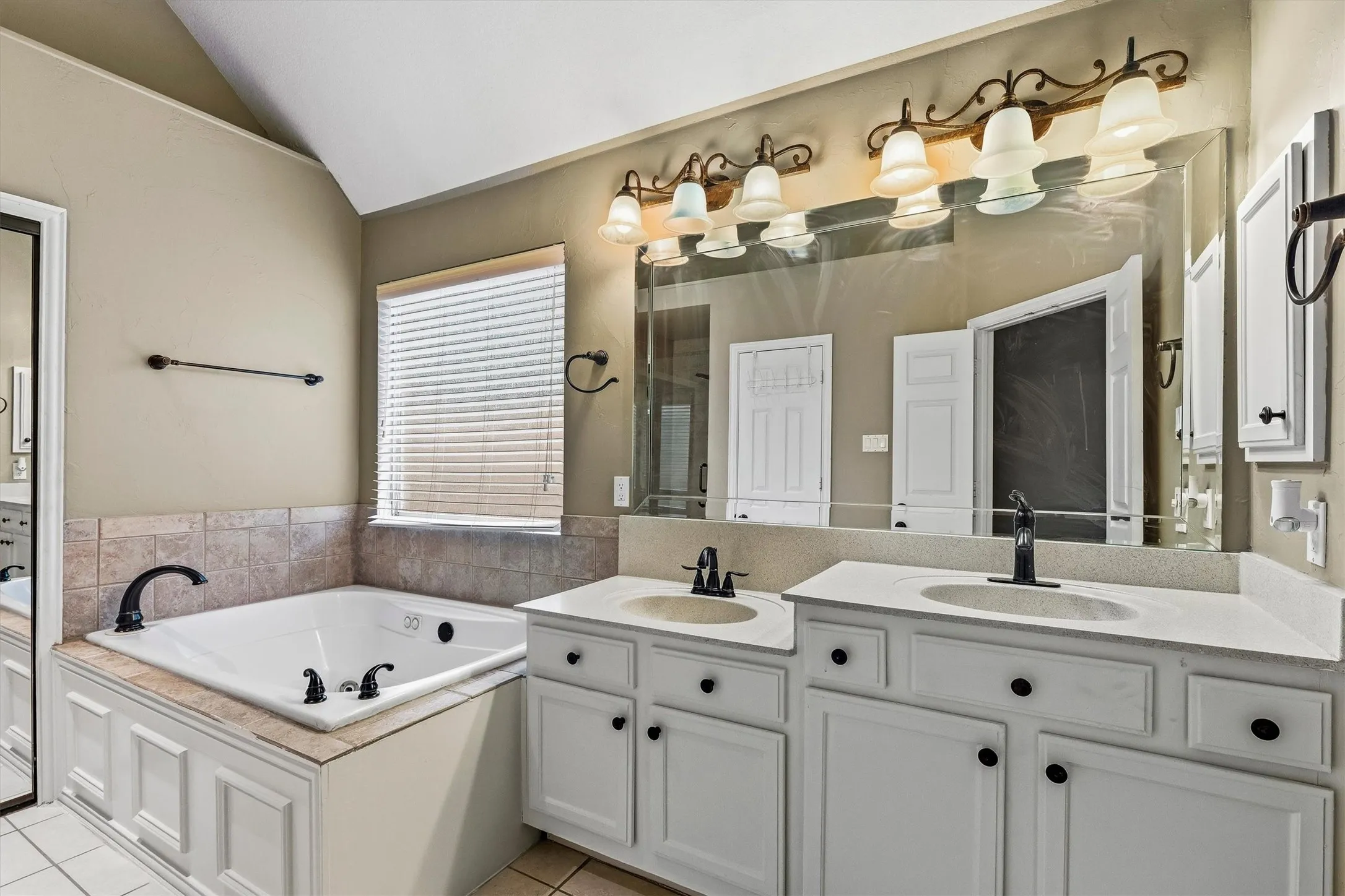Spa-inspired primary bathroom featuring a soaking tub with deck-mounted faucet and fixtures, dual-sink vanity with white cabinetry and brushed nickel hardware, quartz countertops, and a large framed mirror illuminated by a six-light vanity bar