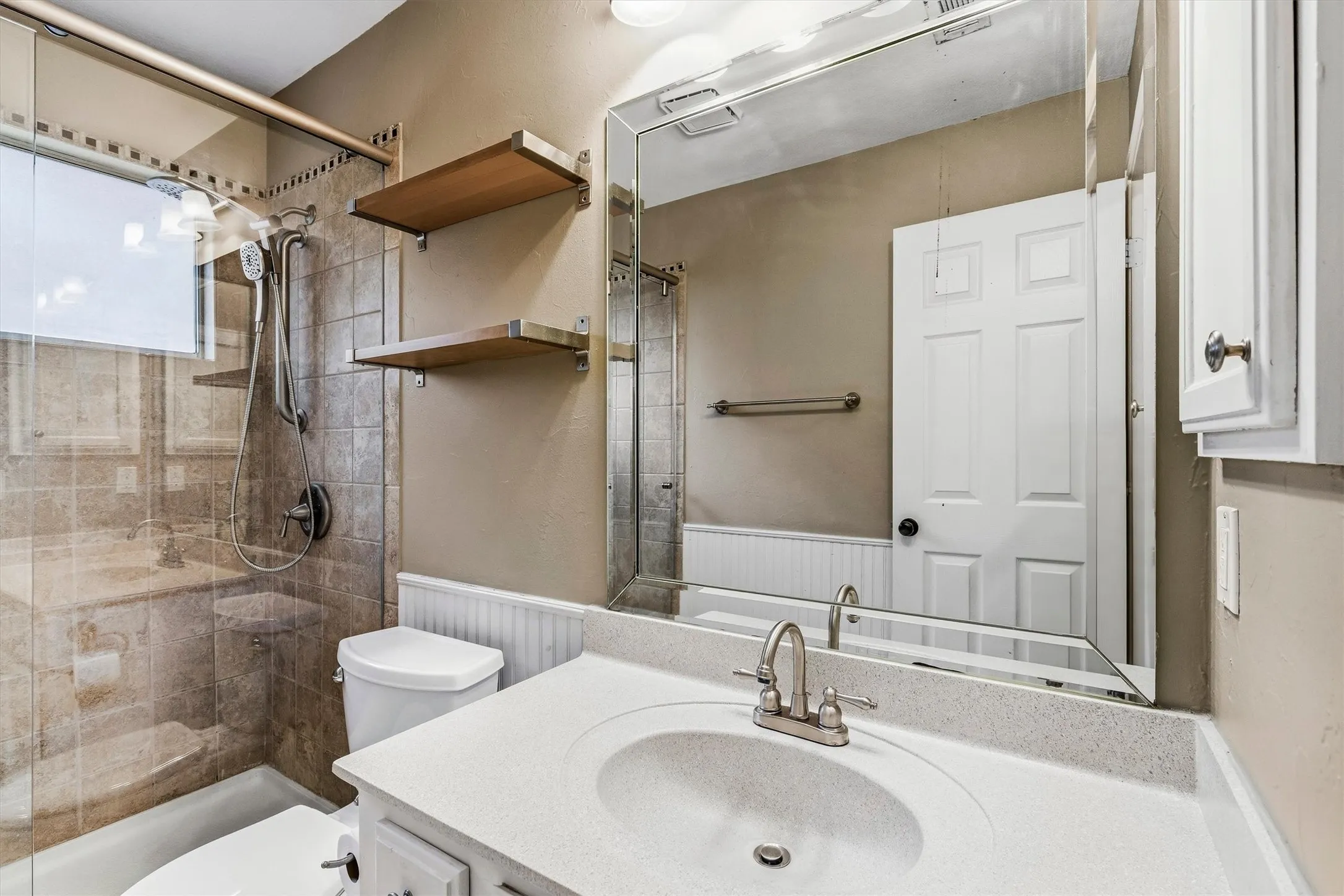 Shower with glass enclosure, white vanity with integrated sink, large wall mirror, and two wood-finish wall shelves