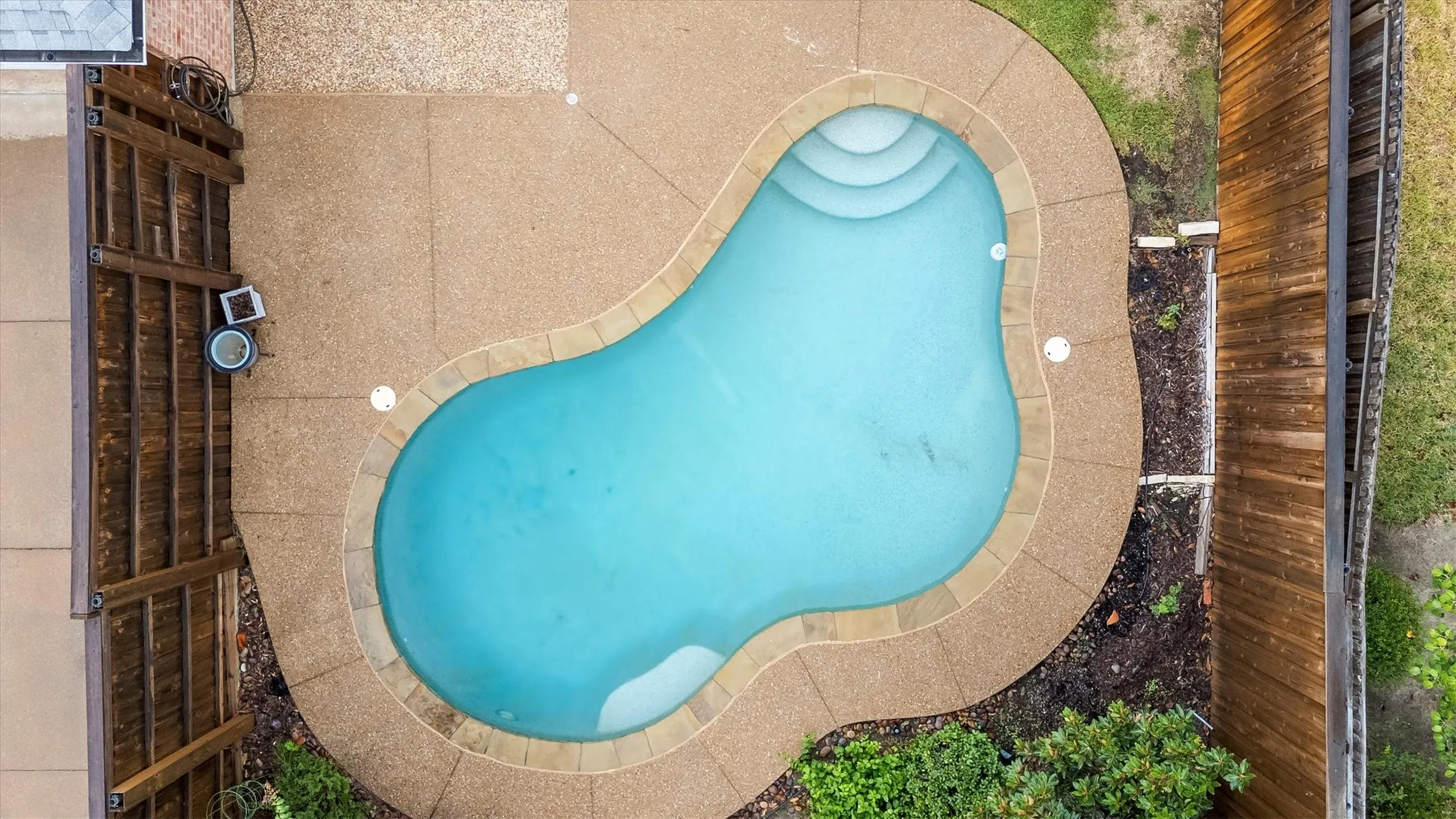 Pear-shaped swimming pool with integrated steps, surrounded by an aggregate concrete patio and perimeter landscaping
