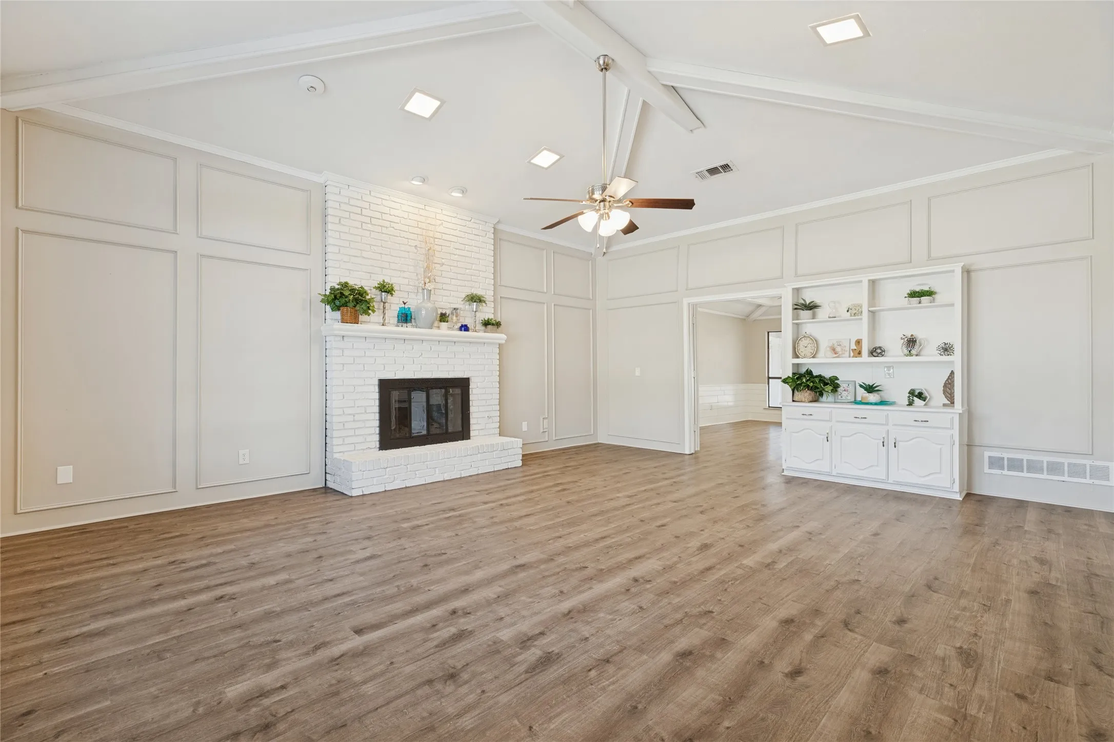 Vaulted ceiling with exposed beams, wood-finish flooring, white brick fireplace, decorative wall paneling, and built in hutch with cabinets
