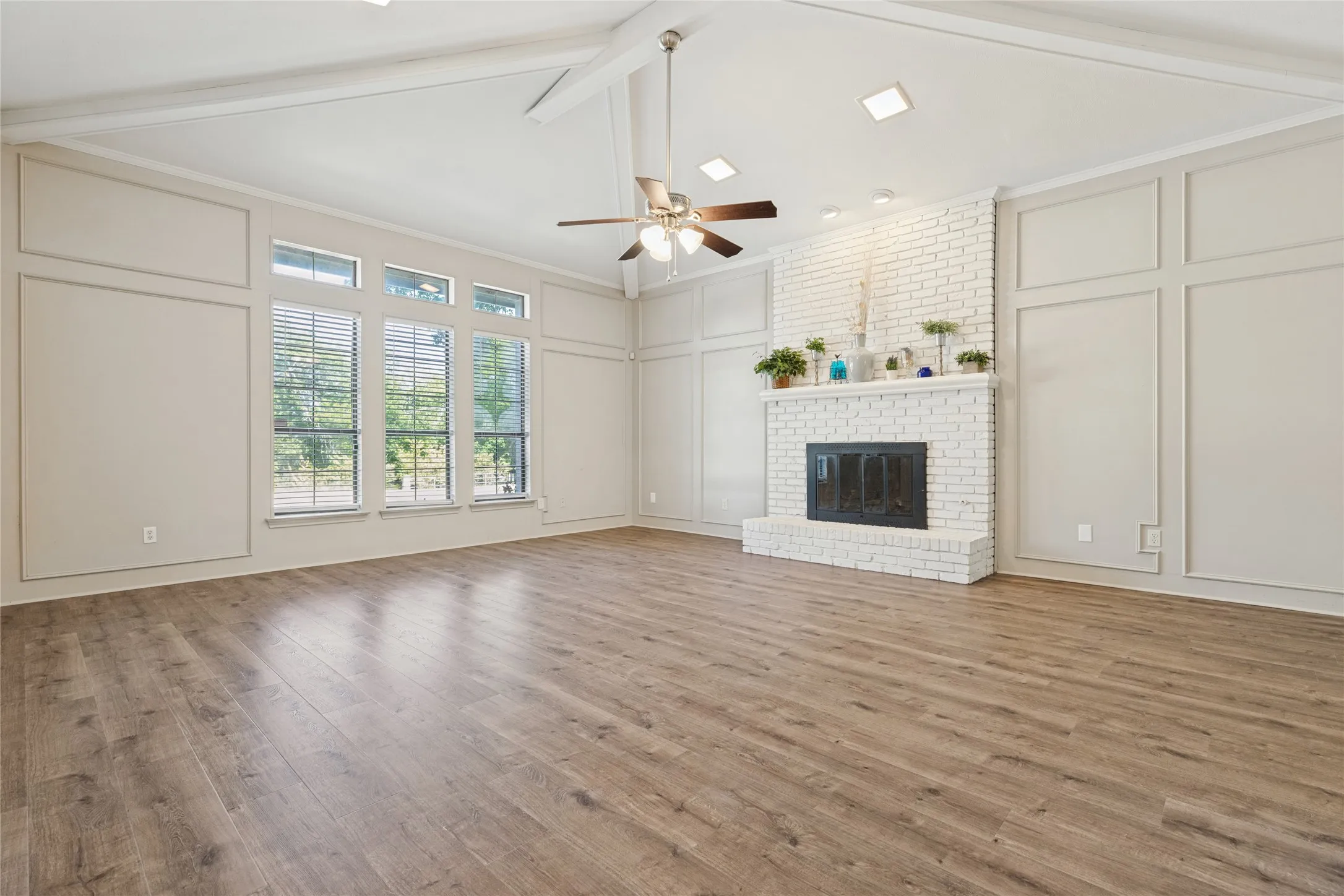 Vaulted ceiling, wood-finish flooring, white brick fireplace with gas starter, picture frame wall paneling, and a ceiling fan with integrated lighting, large windows with park view