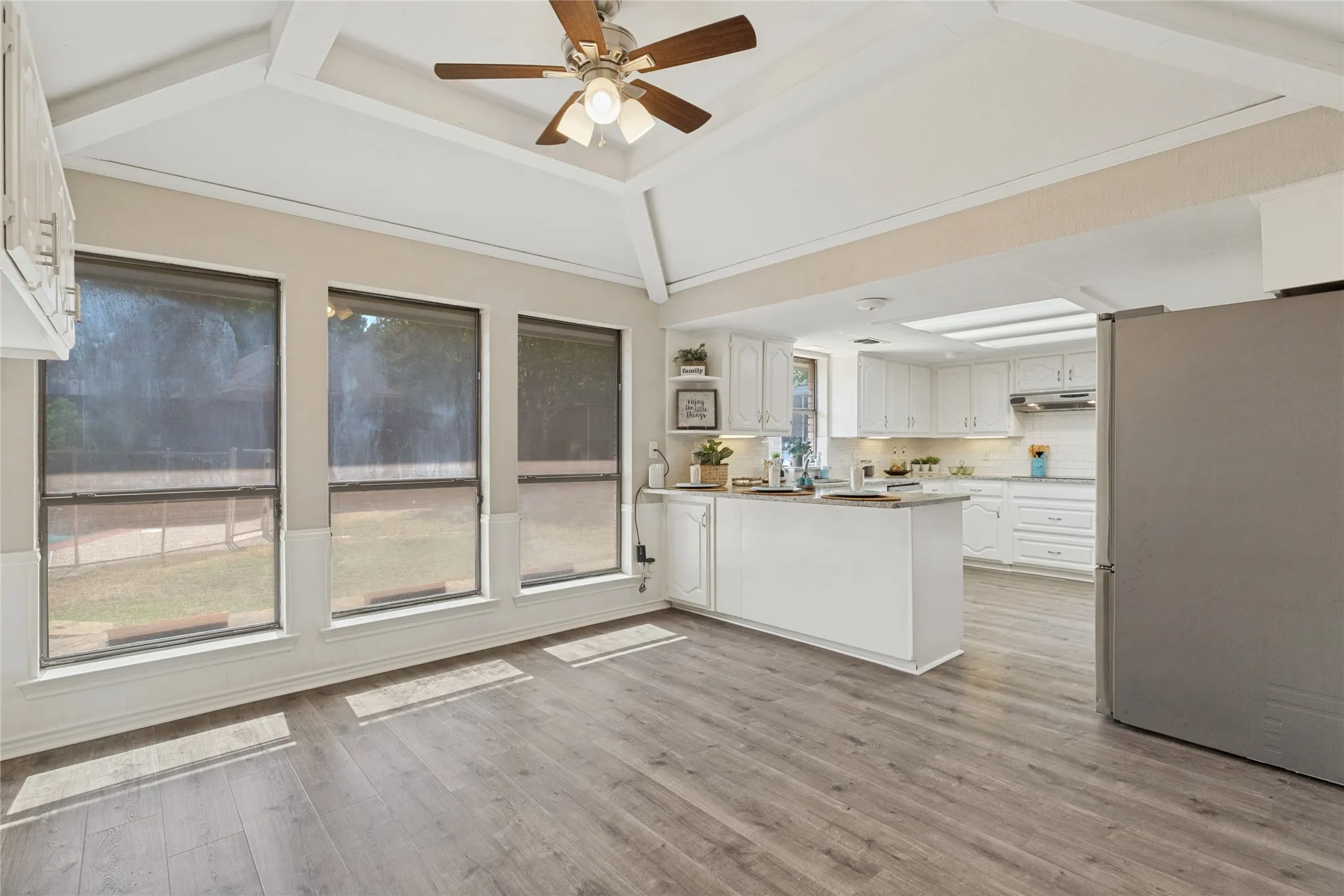Open-concept kitchen and dining area featuring vaulted ceilings with exposed beams, wood-finish flooring, a ceiling fan, and white cabinetry