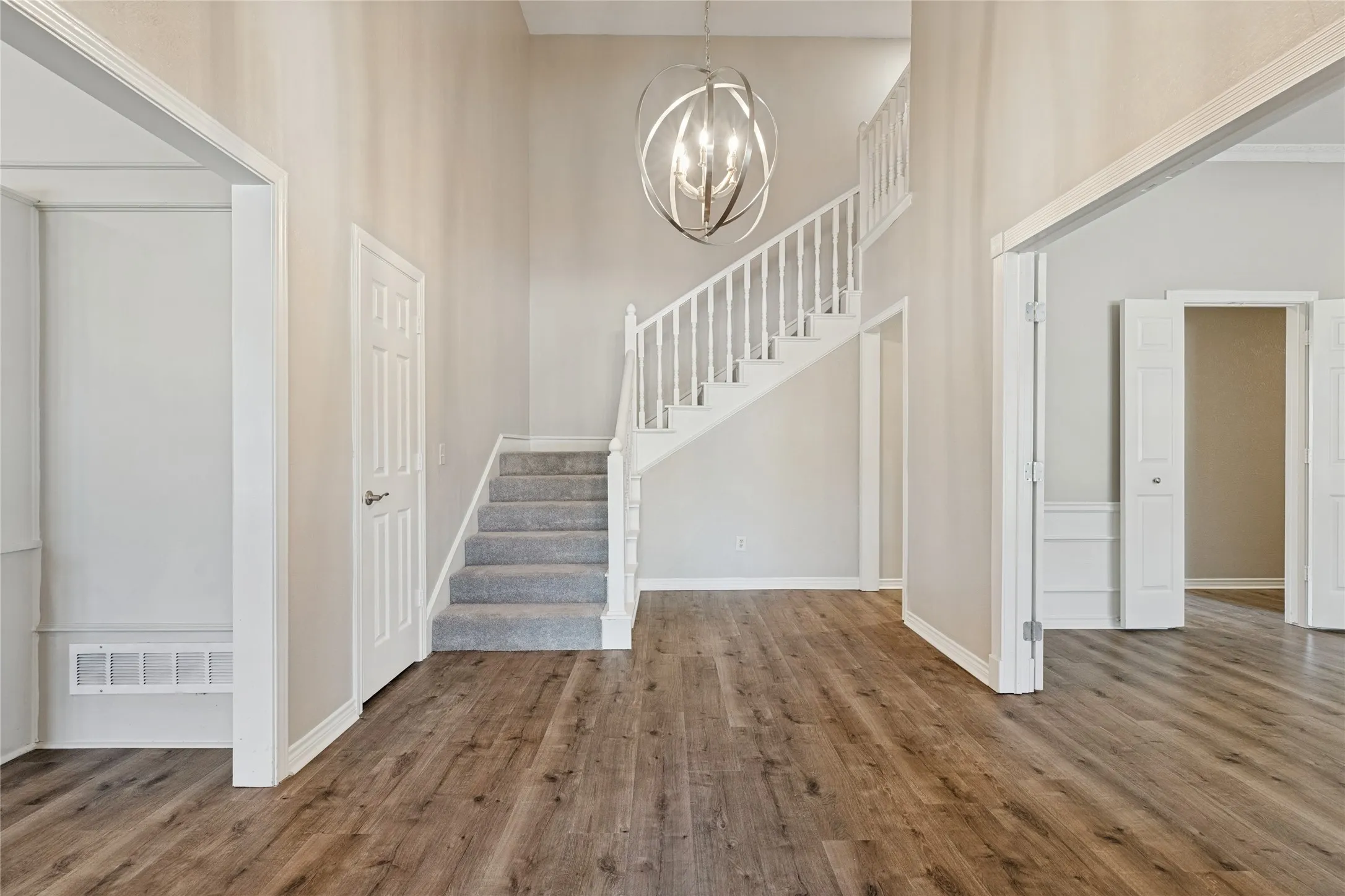 Expansive entryway featuring wood-finish flooring, a contemporary chandelier, a staircase with white balusters and carpeted treads, and paneled interior doors