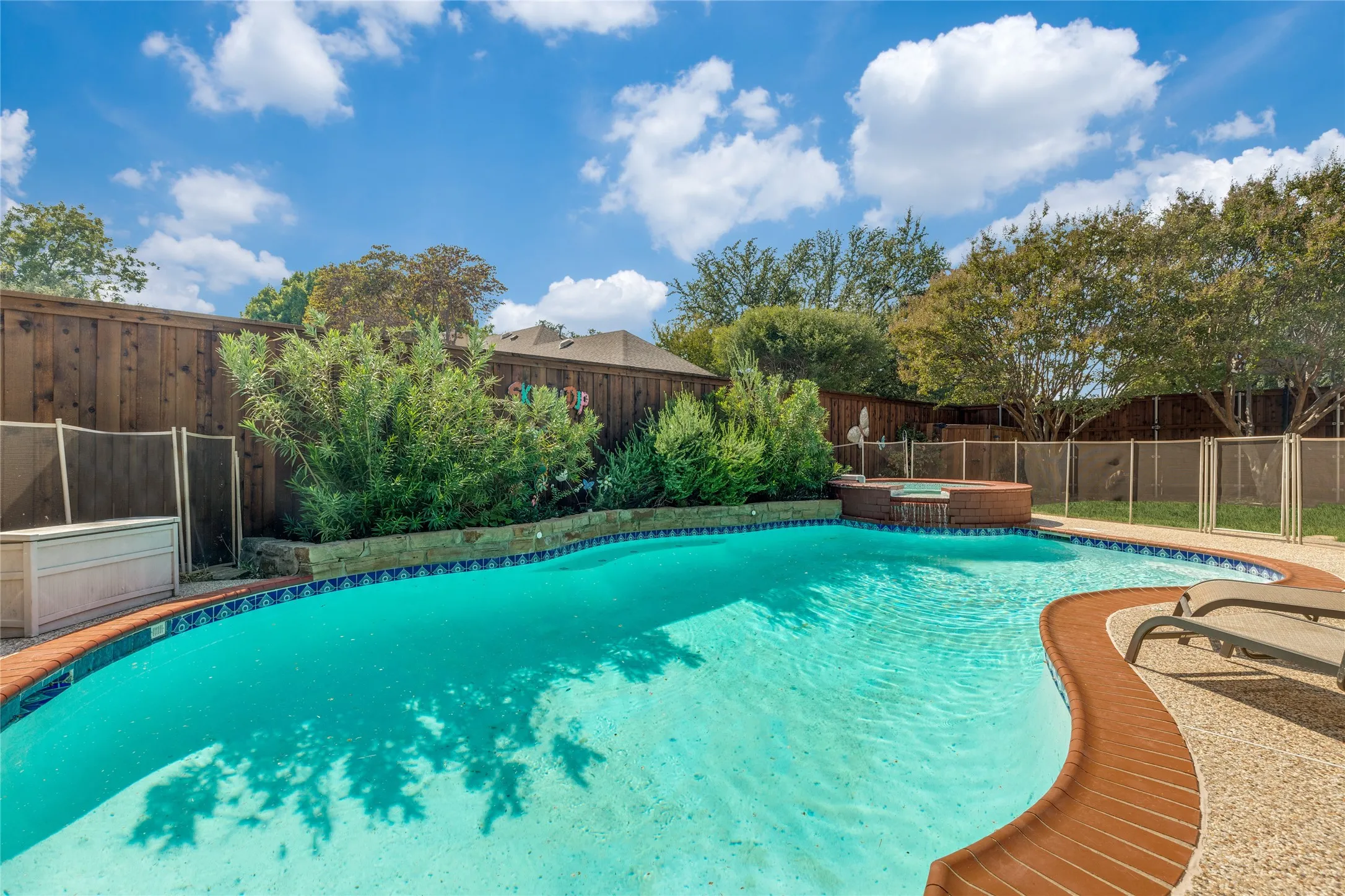 Sparkling swimming pool with a curved design, adjacent hot tub with waterfall feature, stone-edged planting beds, and a wood privacy fence